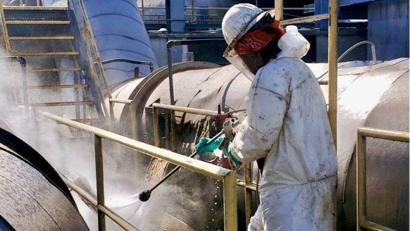 A First Environmental worker  in a protective suit is cleaning a pipe in a factory.