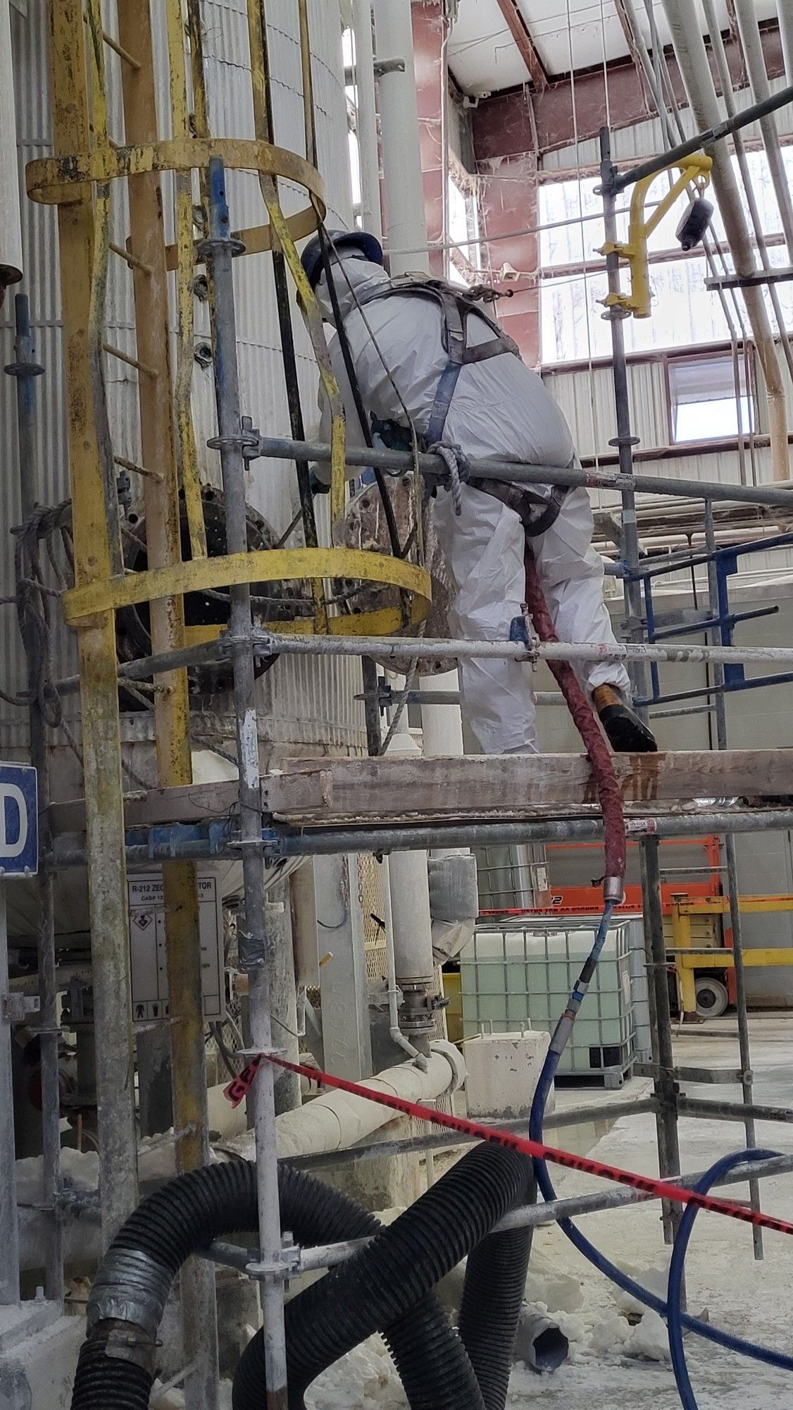 A First Environmental employee in a protective suit is standing on a scaffolding in a factory.