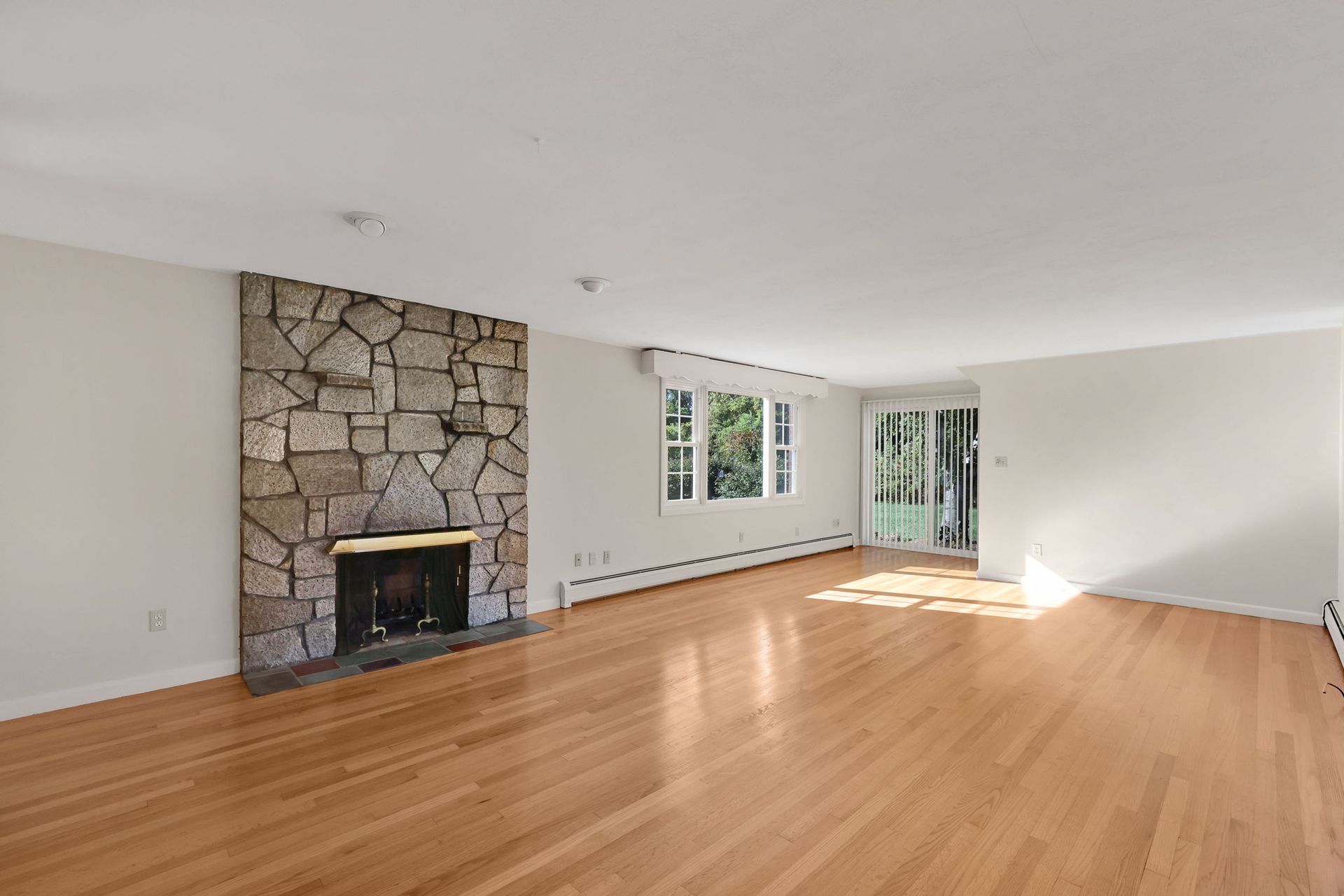 Empty living room with wood floors, stone fireplace, and sliding glass door.