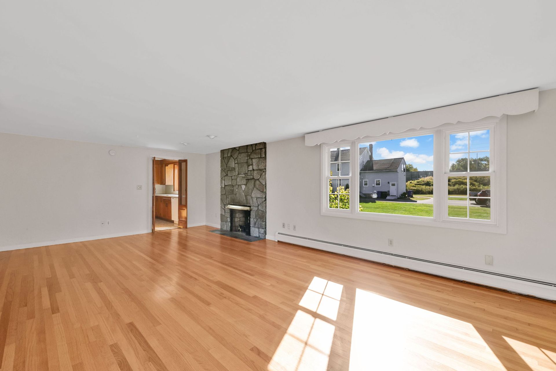 Empty living room with wood floors, stone fireplace, and large window overlooking a yard and small house.