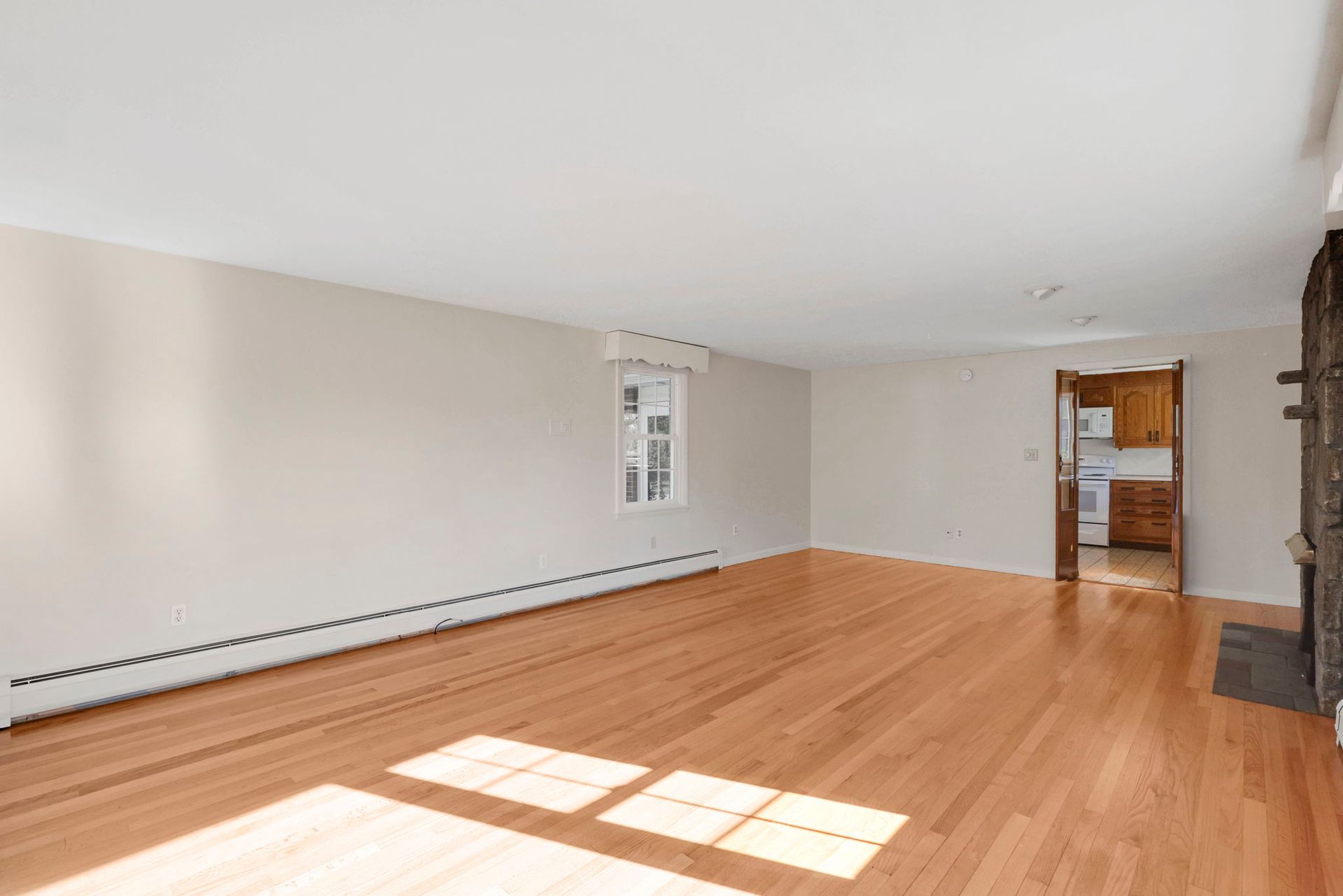 Empty living room with wood floors, light walls, and a glimpse of the kitchen.