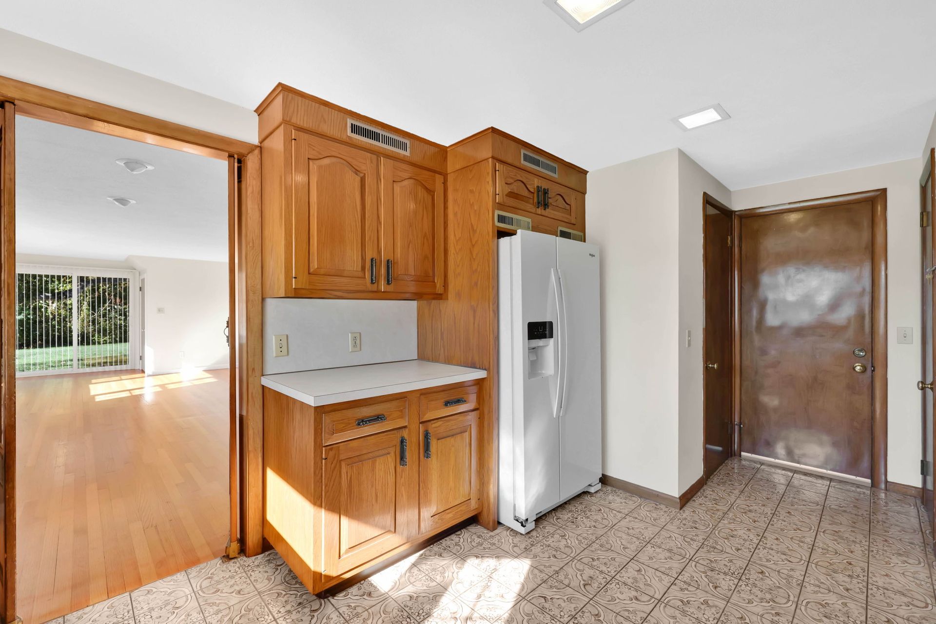 Kitchen with wooden cabinets, refrigerator, and tile floor, door to the living room and entryway.