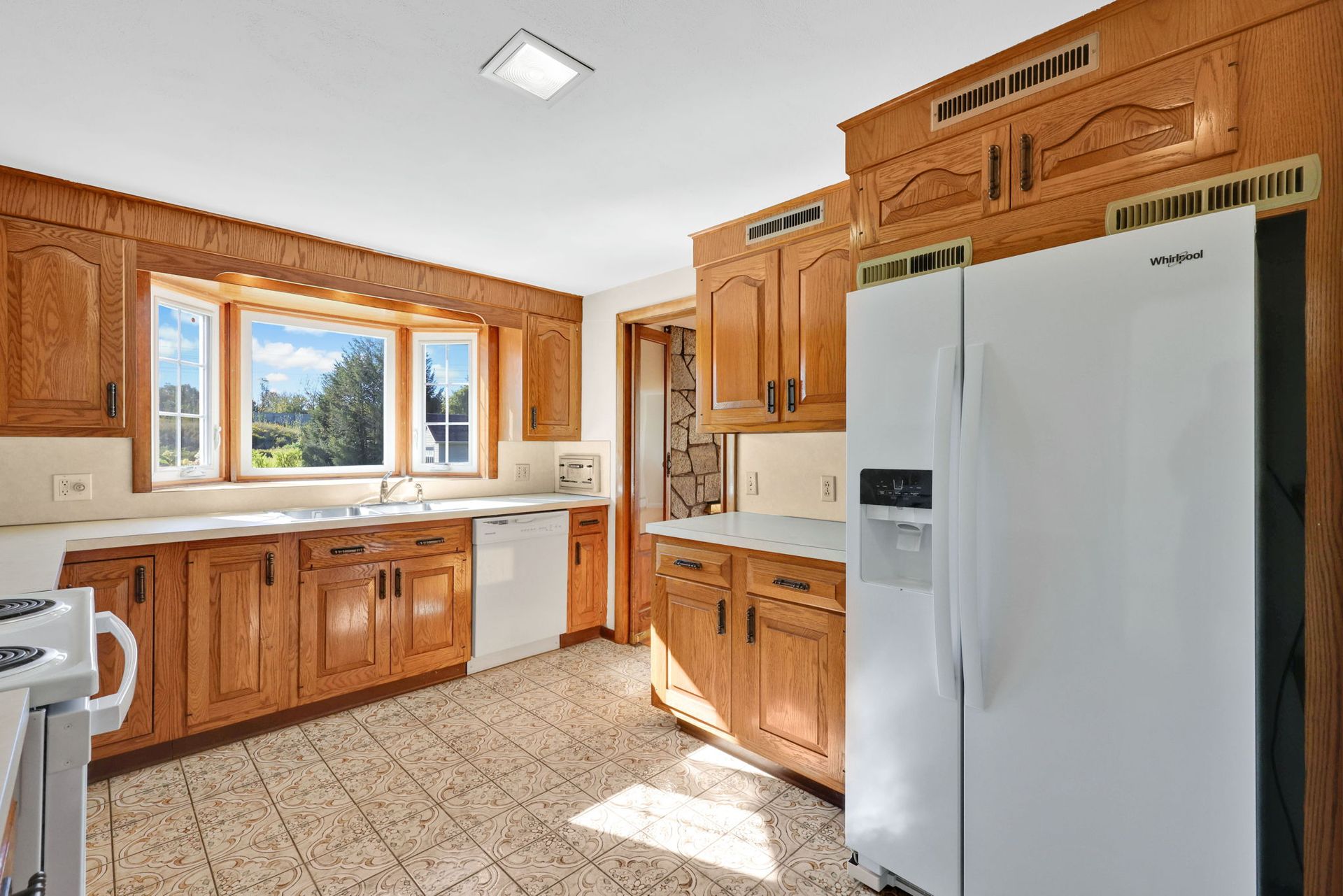 Kitchen with wooden cabinets, white appliances, and a tile floor. A window looks out to greenery.