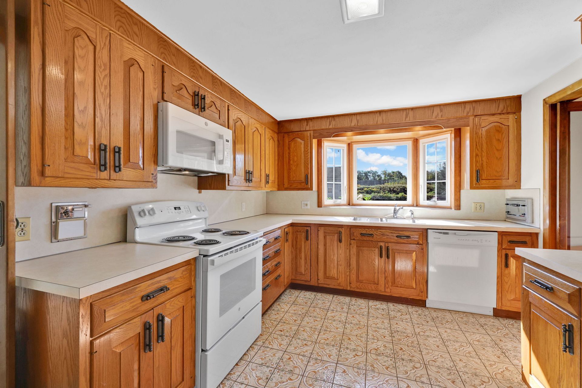 Oak-cabinet kitchen with white appliances and countertops, a bay window, and patterned tile floor.
