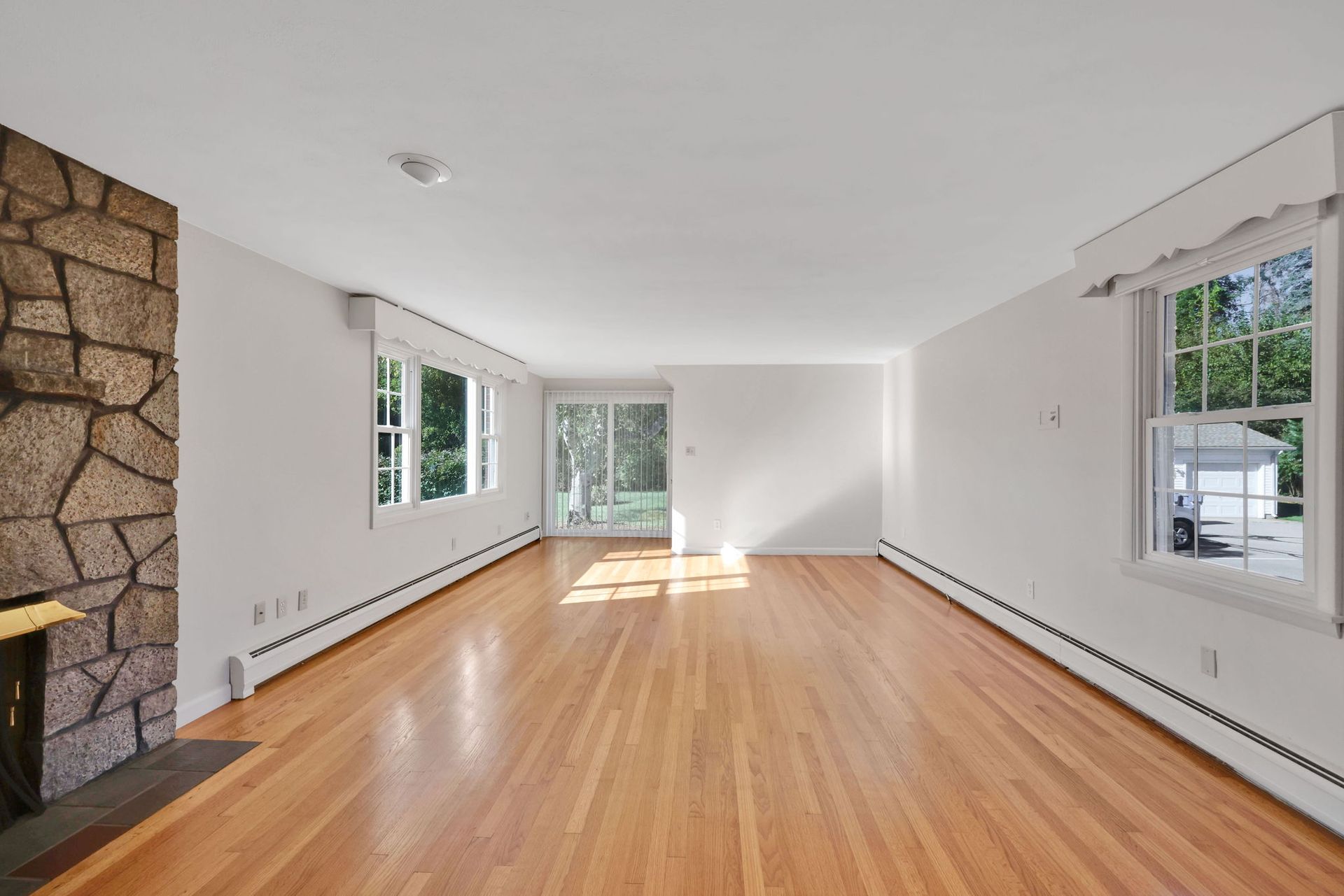 Empty living room with wood floors, stone fireplace, and windows.