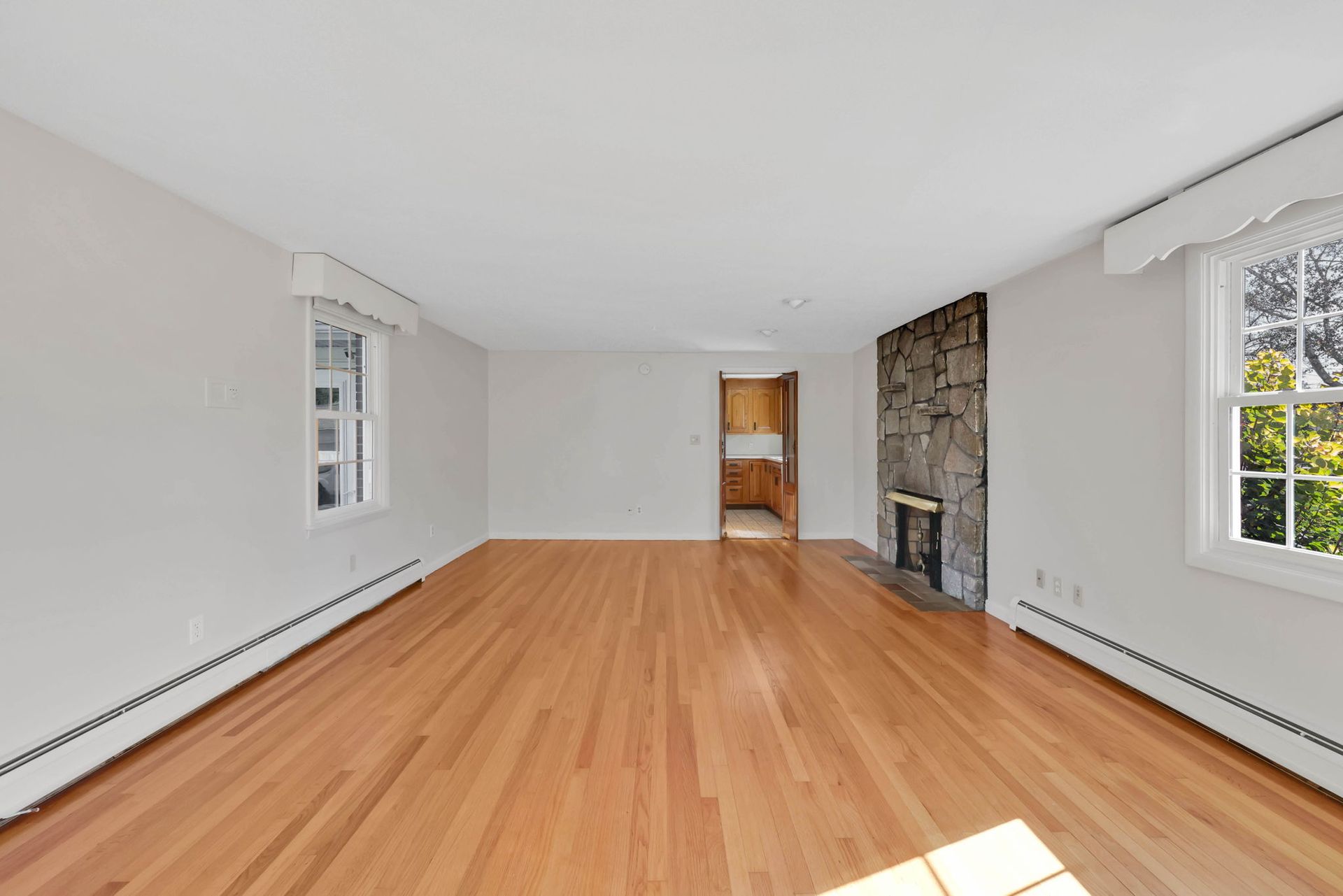 Empty living room with wood floor, stone fireplace, and two windows.