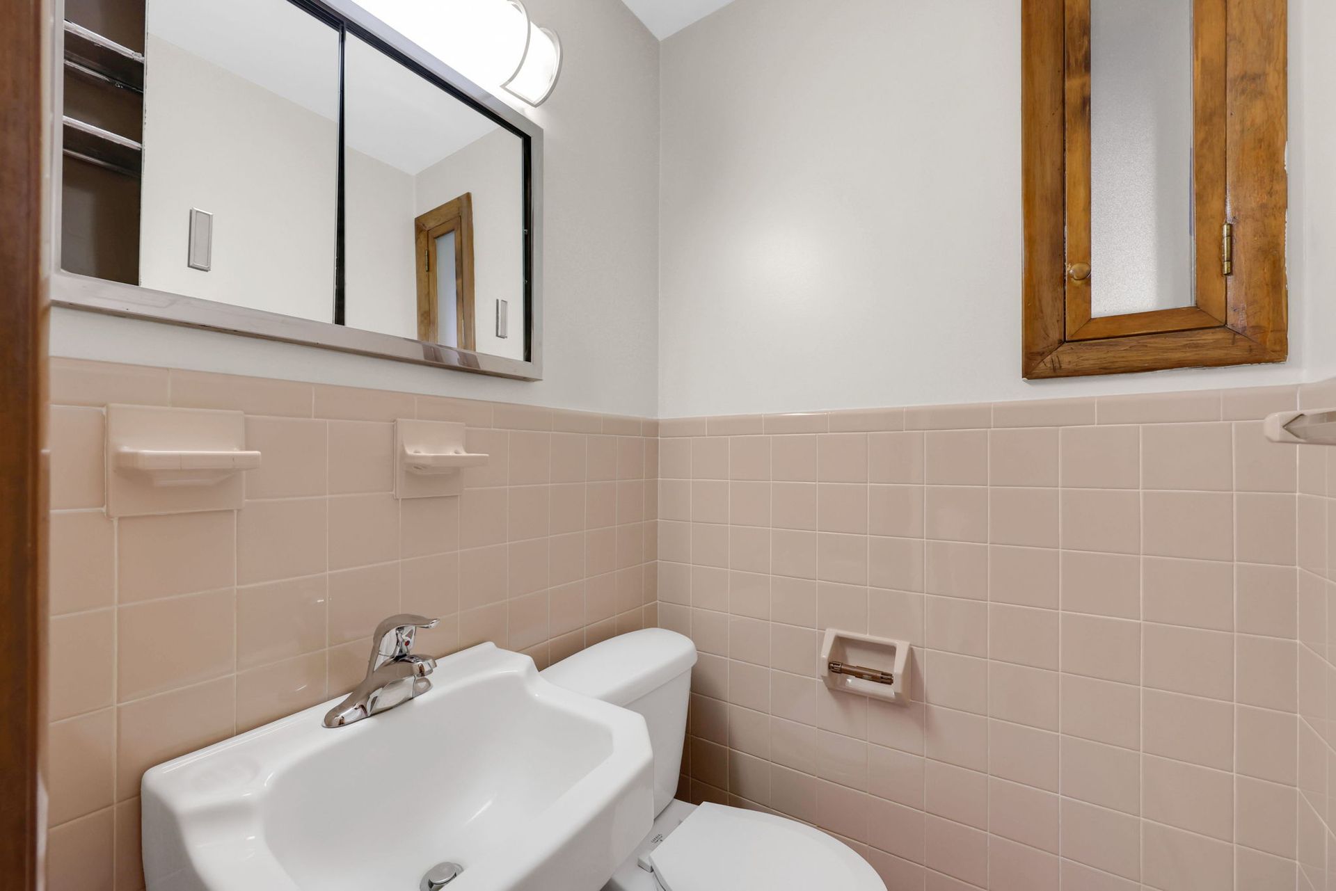 Small bathroom with pink tiled walls, toilet, sink, and two mirrors.
