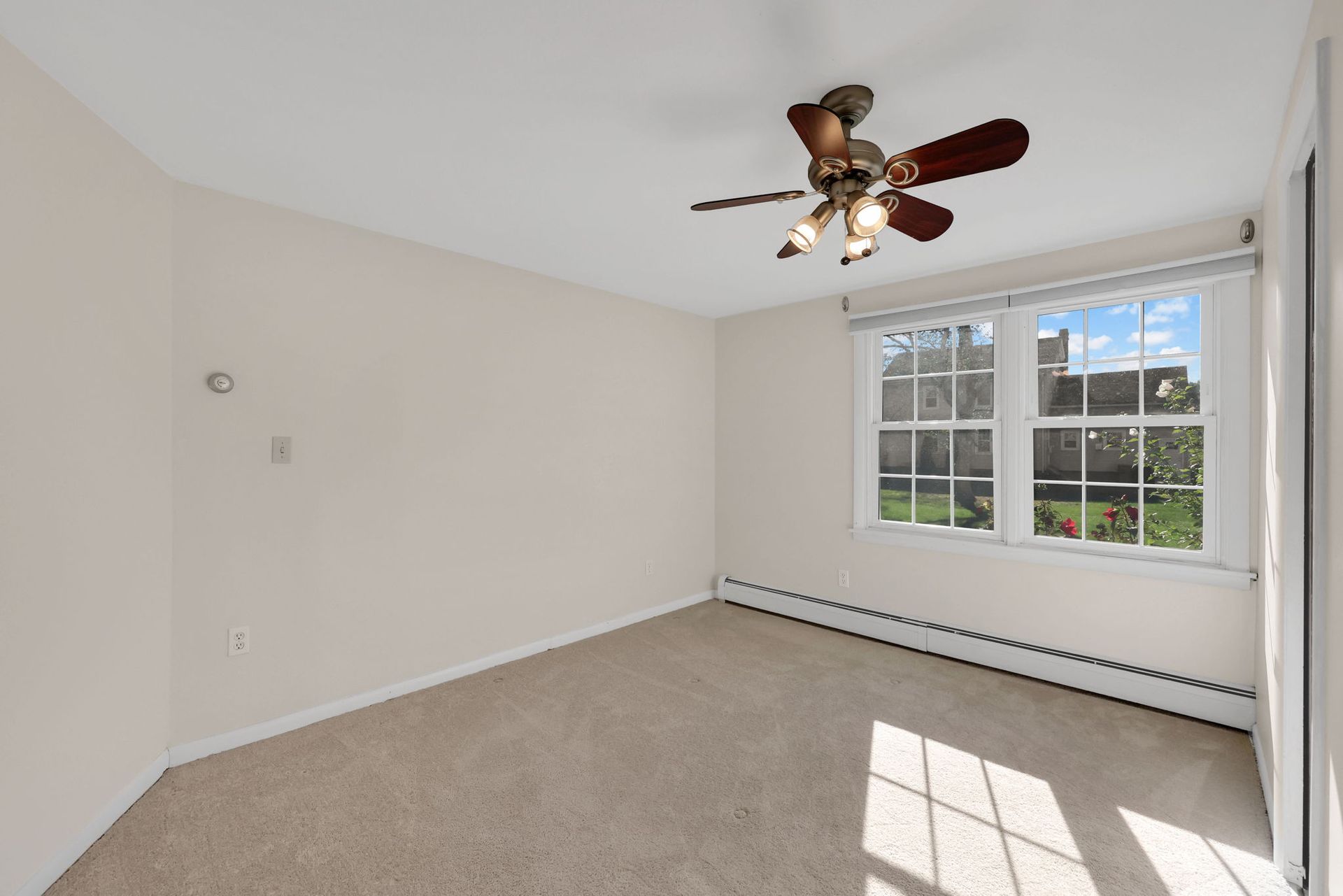 Empty room with beige walls, carpet, and a ceiling fan; a window with sunlight.