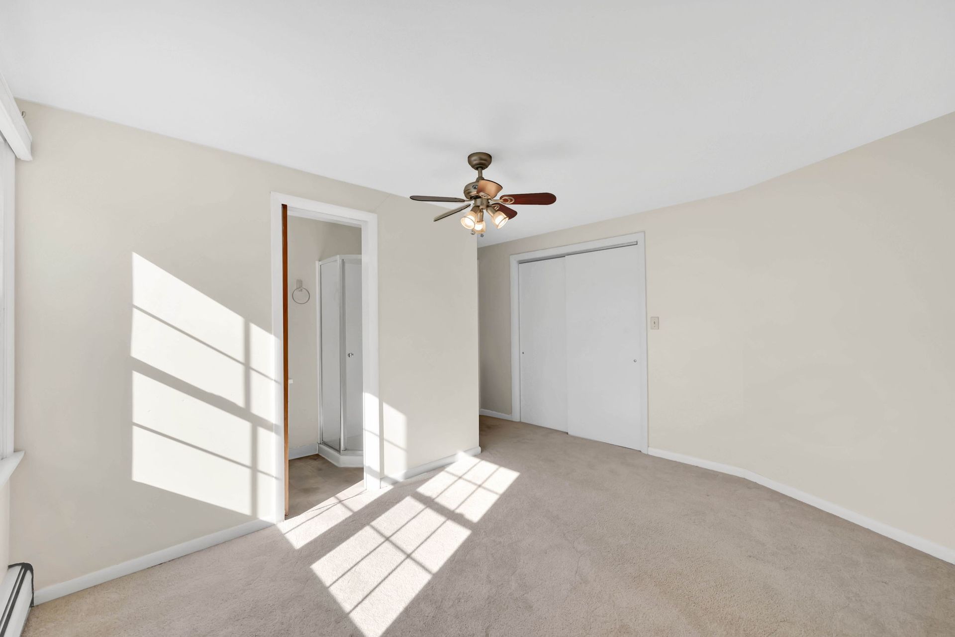 Empty bedroom with carpet, a ceiling fan, a closet, and a bathroom doorway. Sunlight streams in.