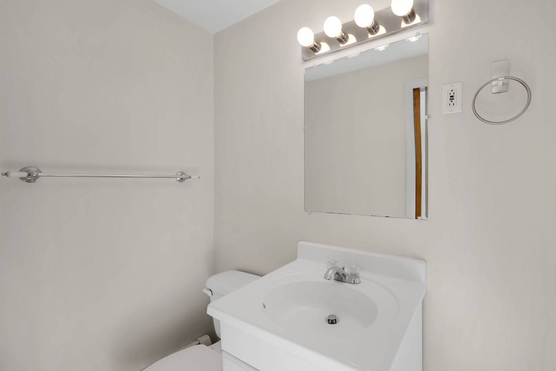 A small, empty bathroom with a sink, mirror, toilet, and towel rack. Light walls, neutral tones.
