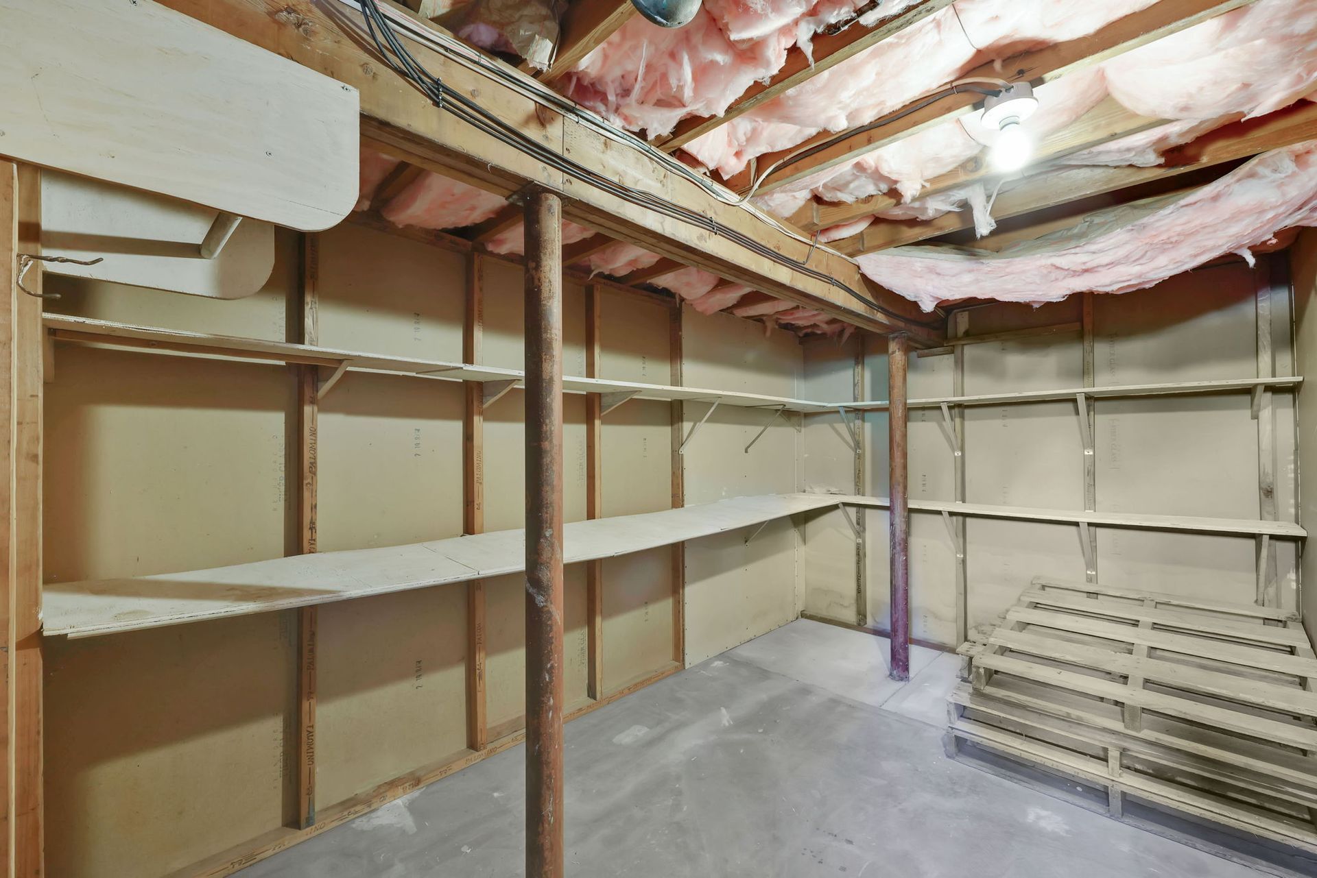 Basement storage room with shelves, support beams, pink insulation, and concrete floor.