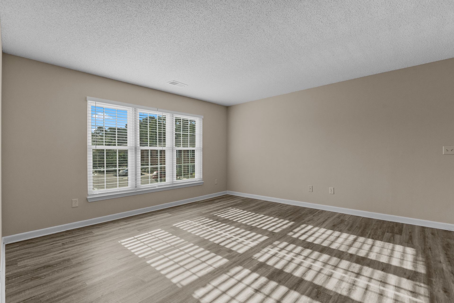 Empty room with a window letting in light, casting striped shadows on the wood floor and neutral-colored walls.