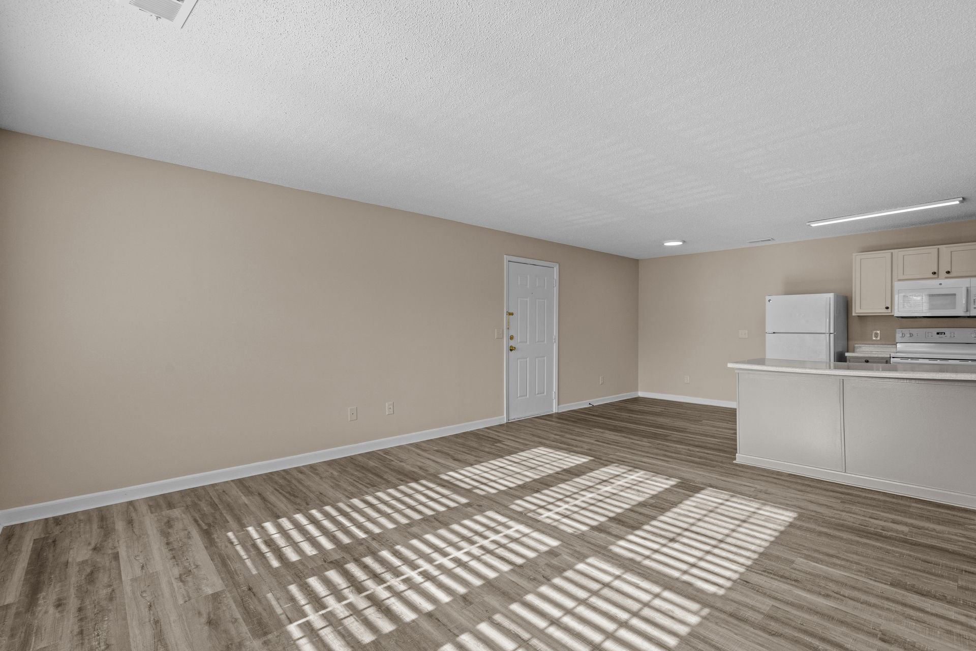 Empty apartment interior with neutral walls, wood-look floors, and kitchen area.