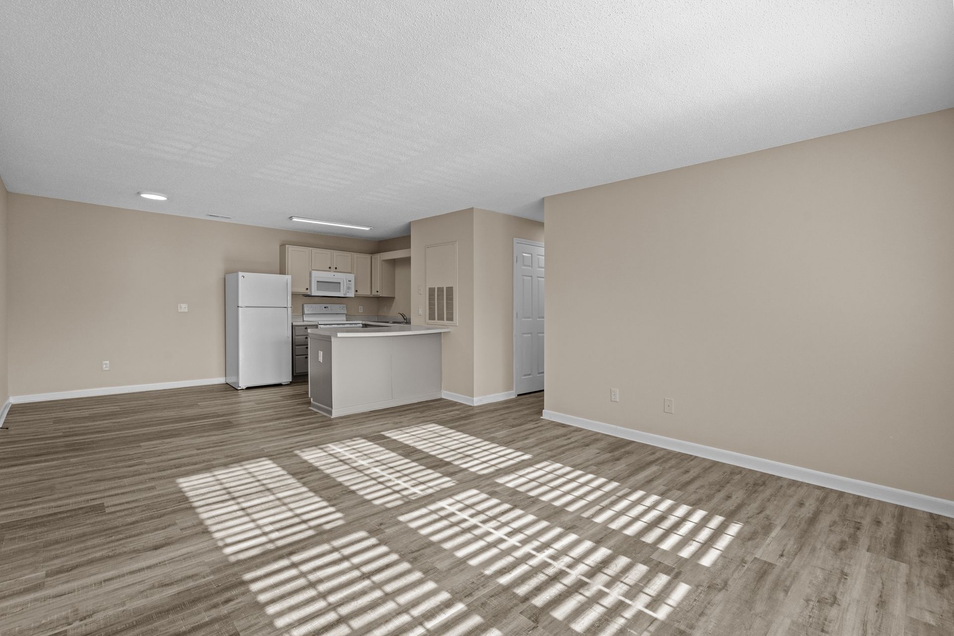 Empty apartment interior with kitchen, laminate flooring, and blinds casting shadows.