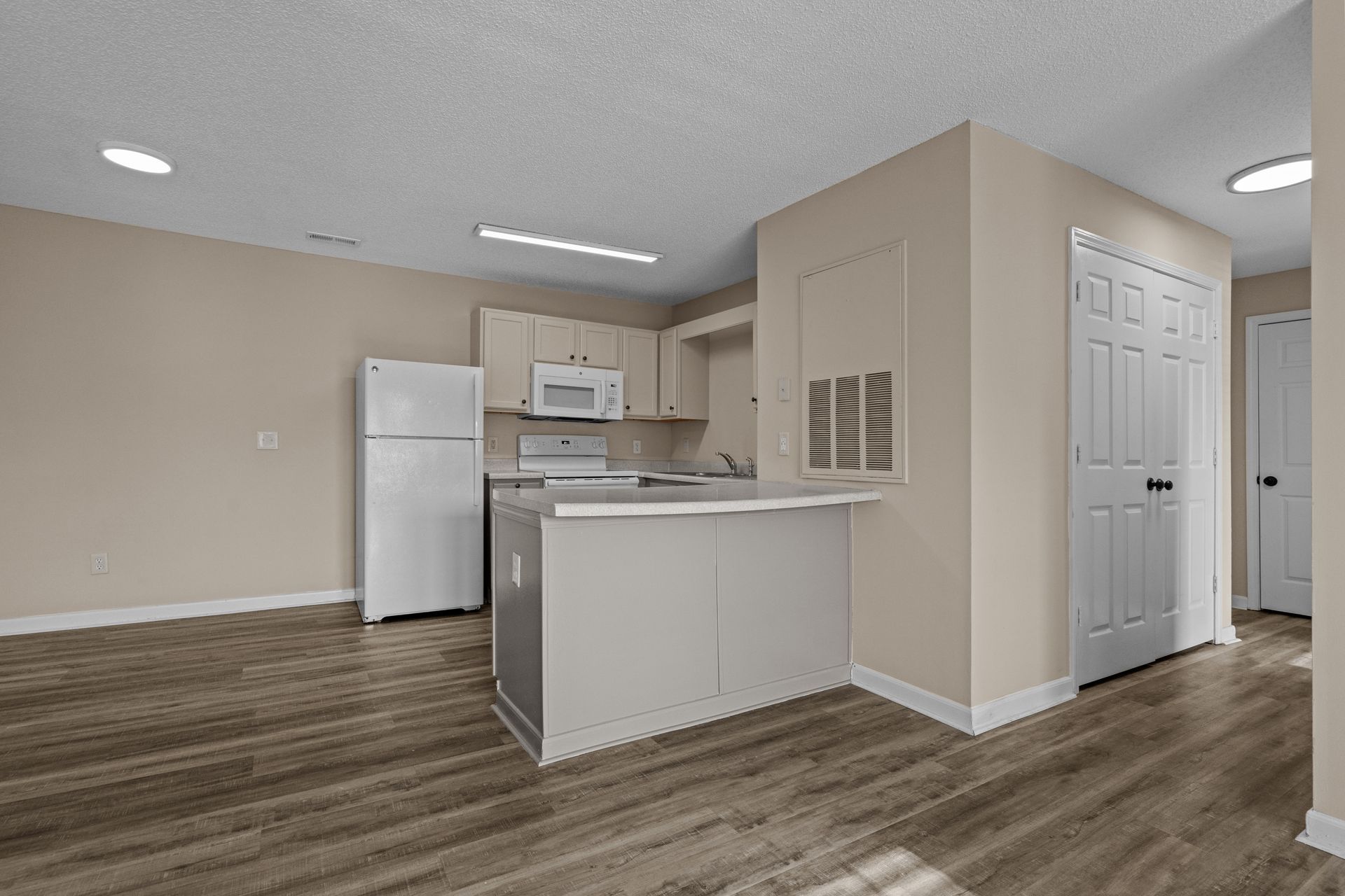 Empty, neutral-toned apartment interior with a kitchen, featuring white appliances, a breakfast bar, and dark wood-look flooring.
