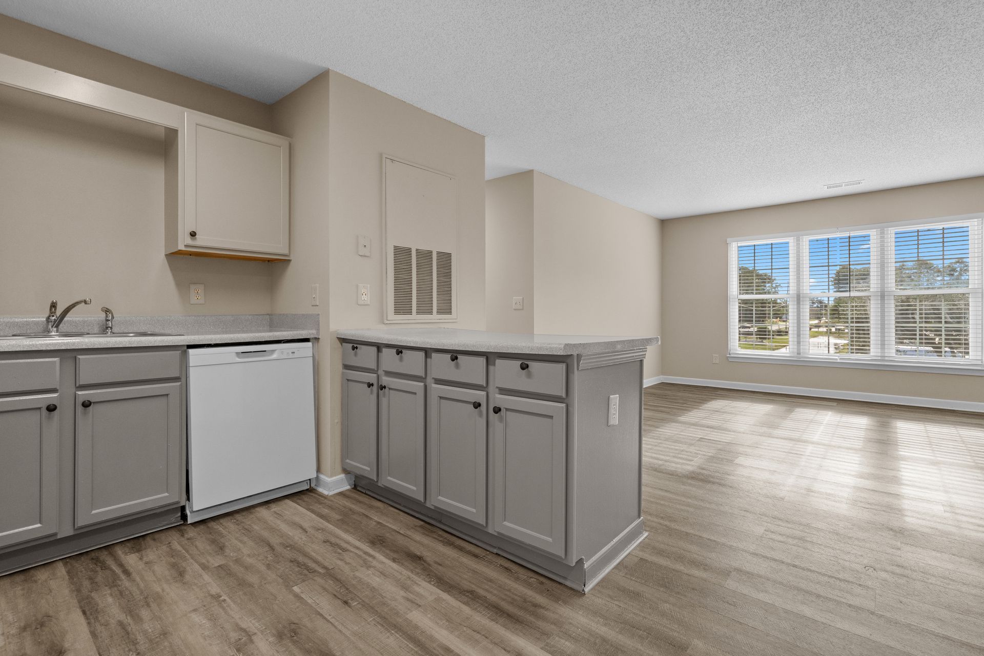 Kitchen with gray cabinets, white appliances, and a countertop island next to a sunny living room with windows.