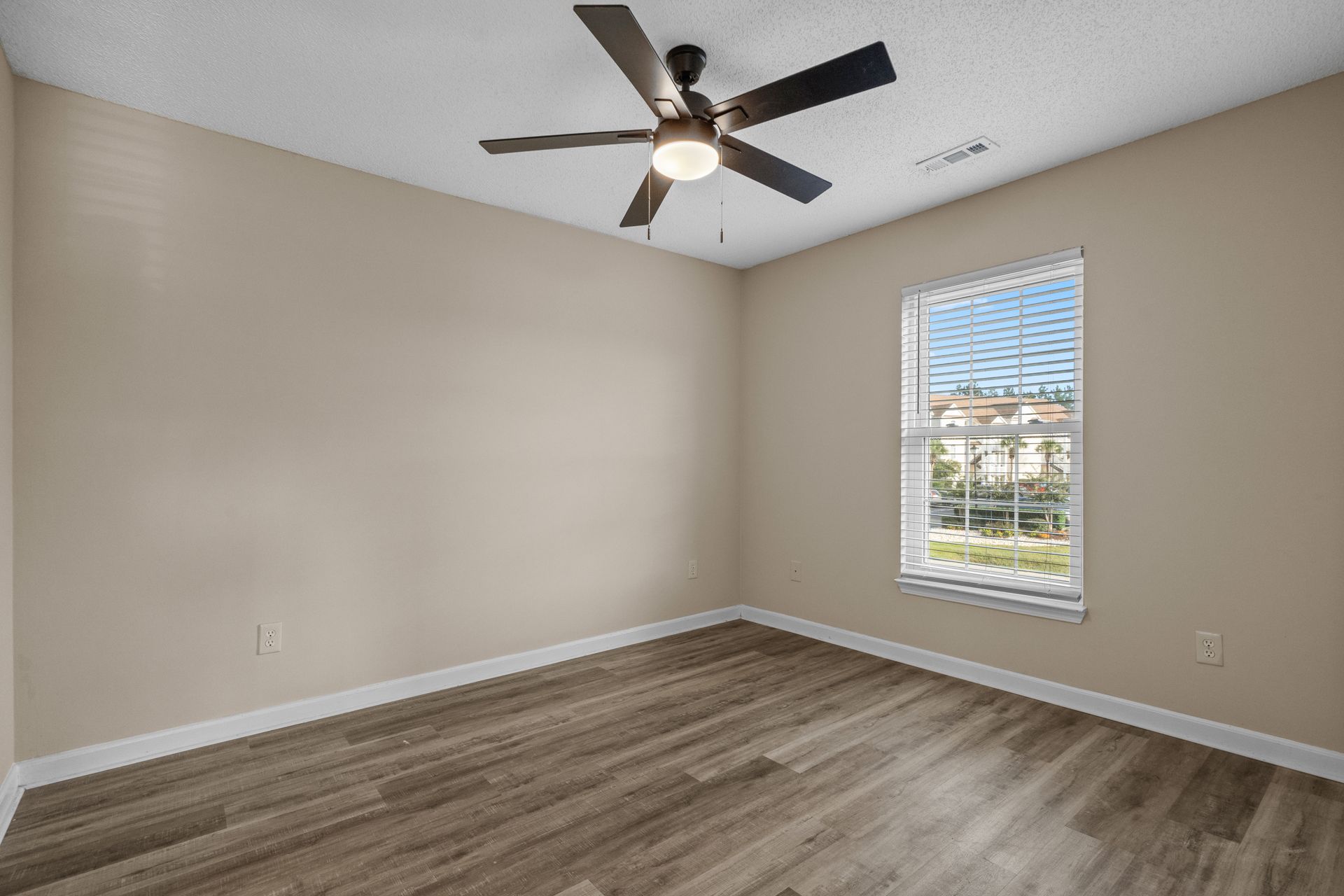 Empty bedroom with tan walls, wood floors, ceiling fan, and window.