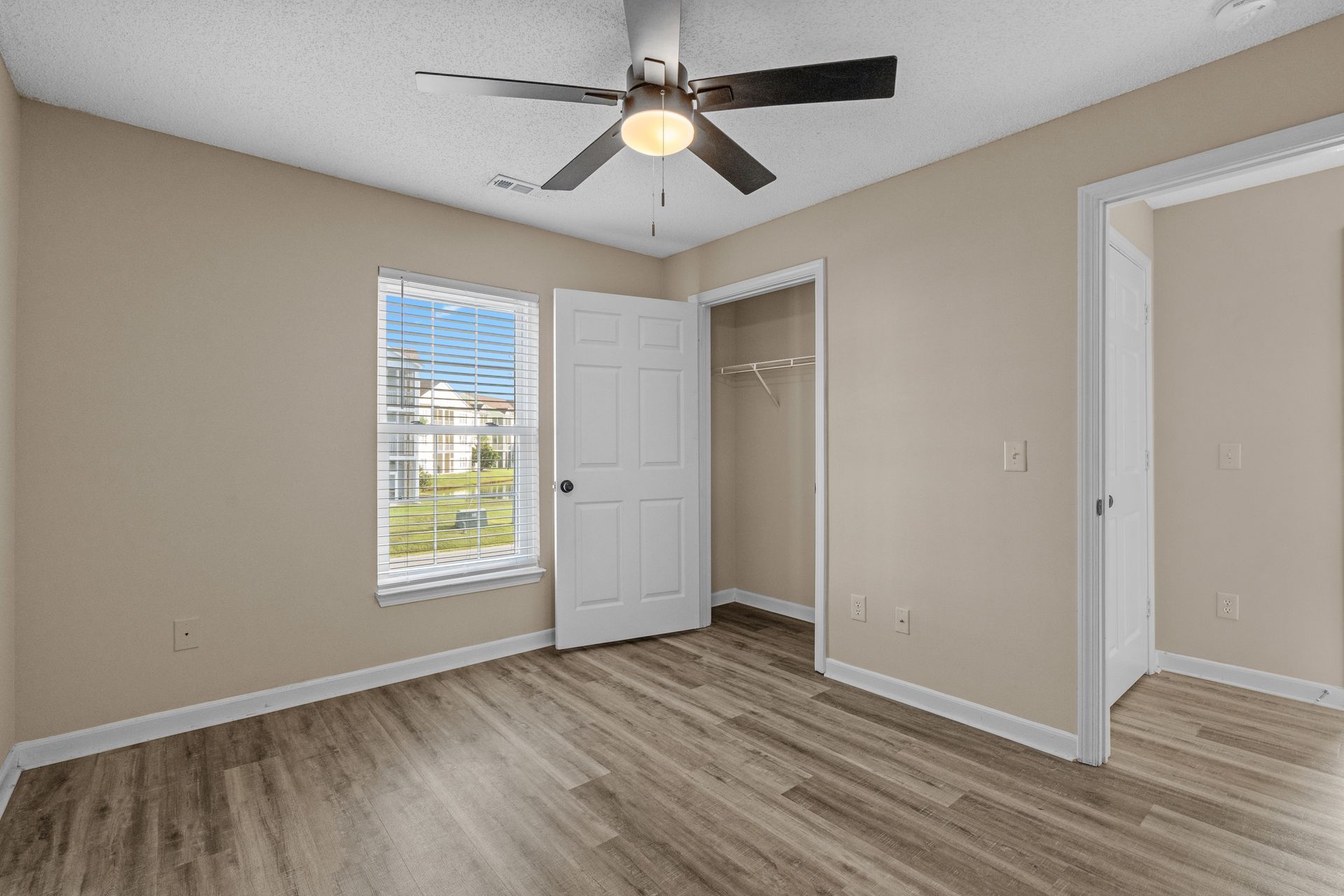 Bedroom interior with beige walls, wood-look flooring, window, closet, ceiling fan, and open door.