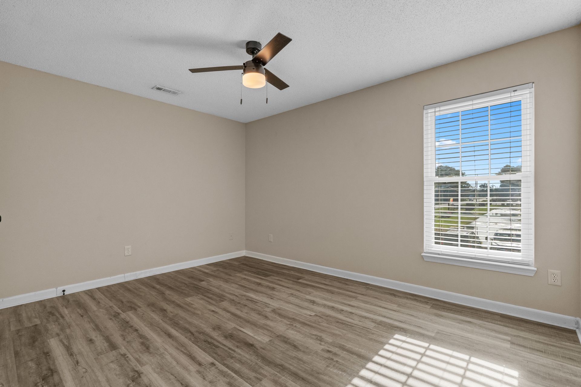Empty bedroom with beige walls, wood-look flooring, ceiling fan, and window with sunlight.