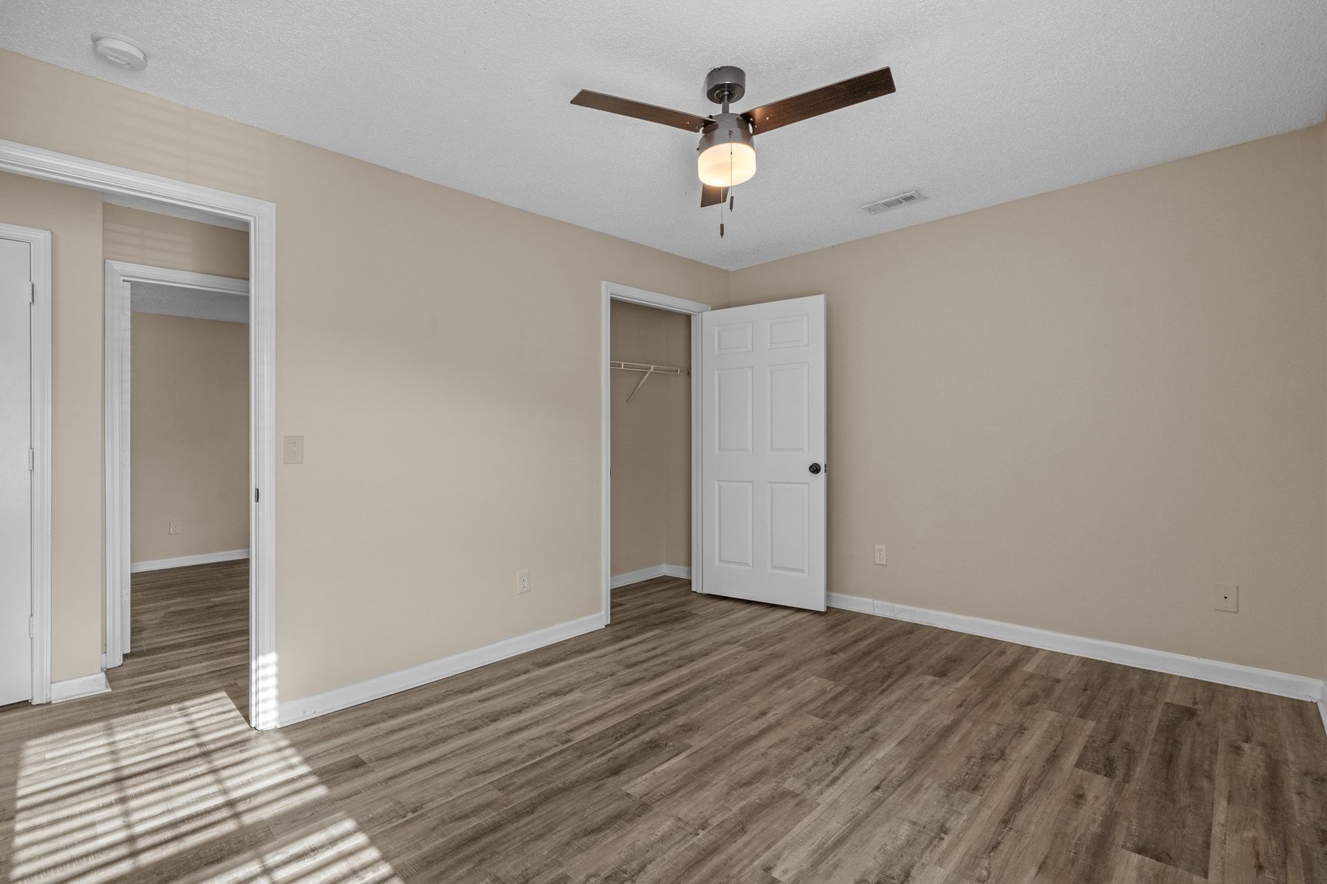 Empty bedroom with beige walls, wood-look flooring, ceiling fan, and open doors to closets and another room.