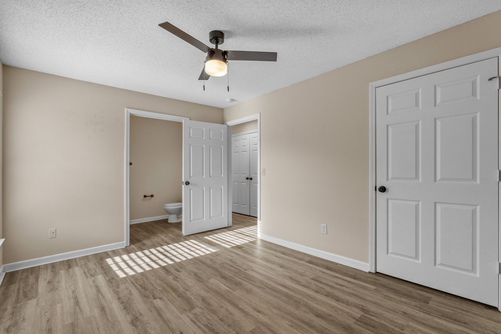 Bedroom with wooden floor, white doors, beige walls, and a ceiling fan. Two doors open into other rooms.