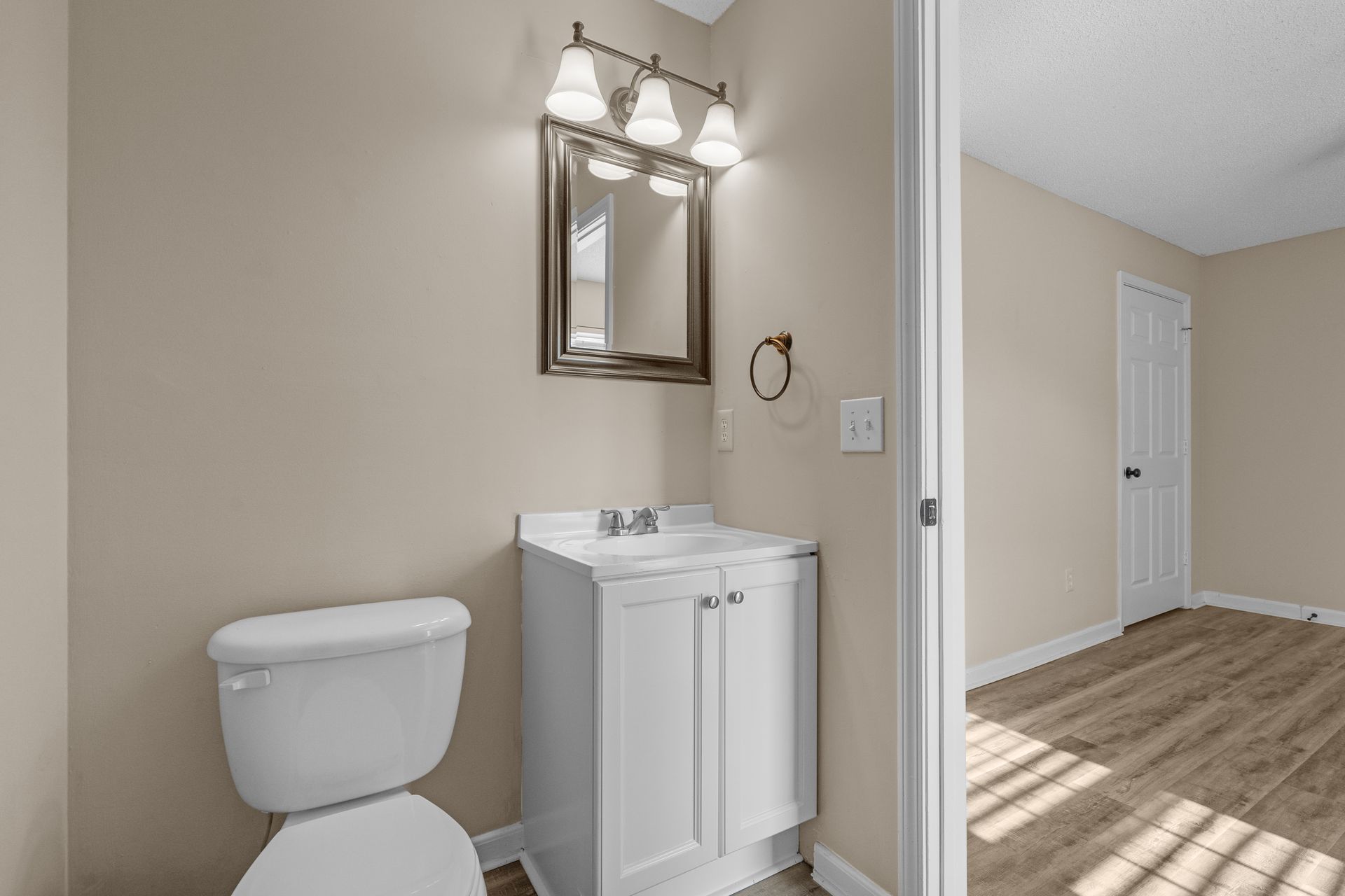 A small bathroom with a toilet, vanity, and open doorway to a room with wood-look flooring.