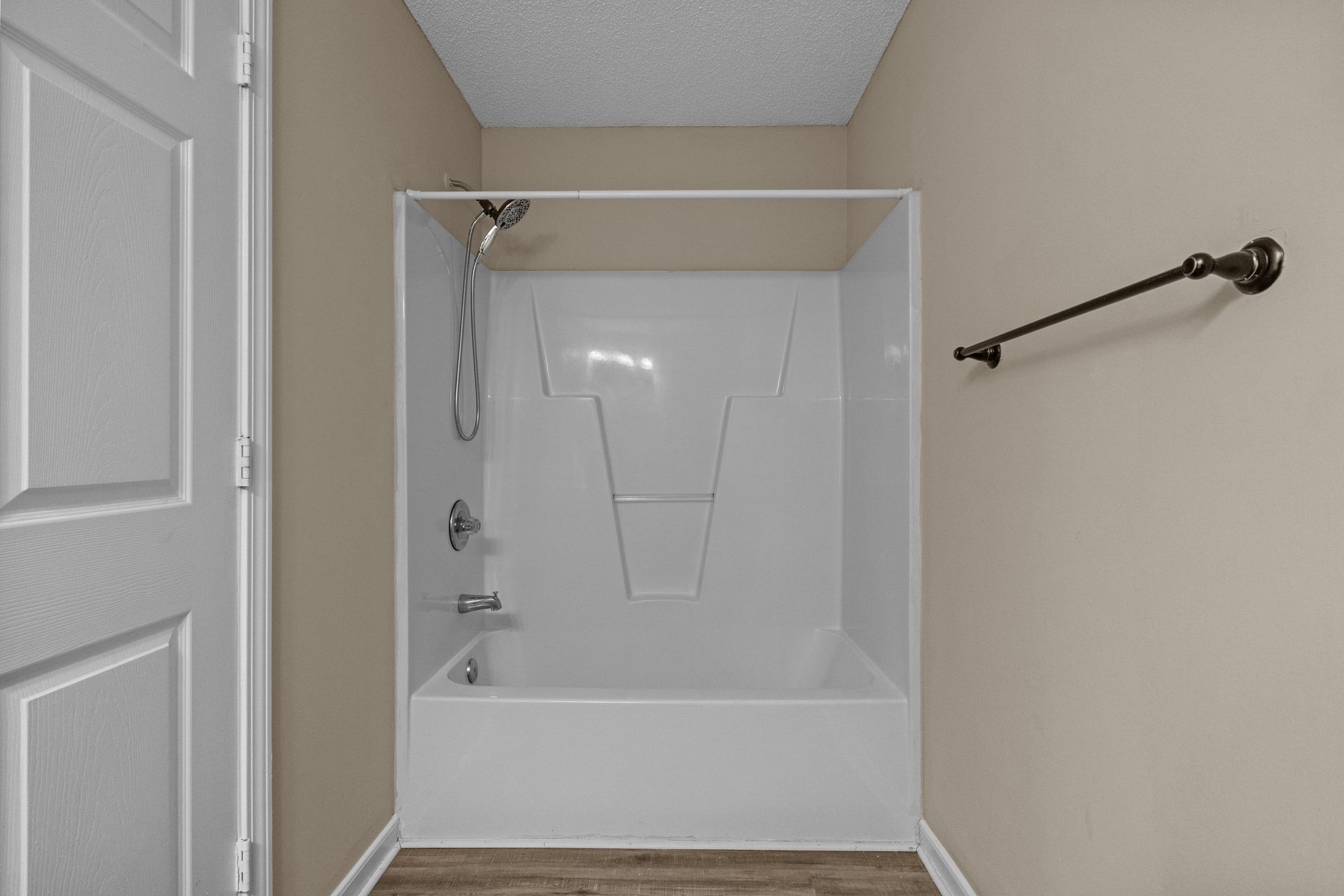 Bathroom with white shower and tan walls. Brown towel bar on the right. Wooden floor.