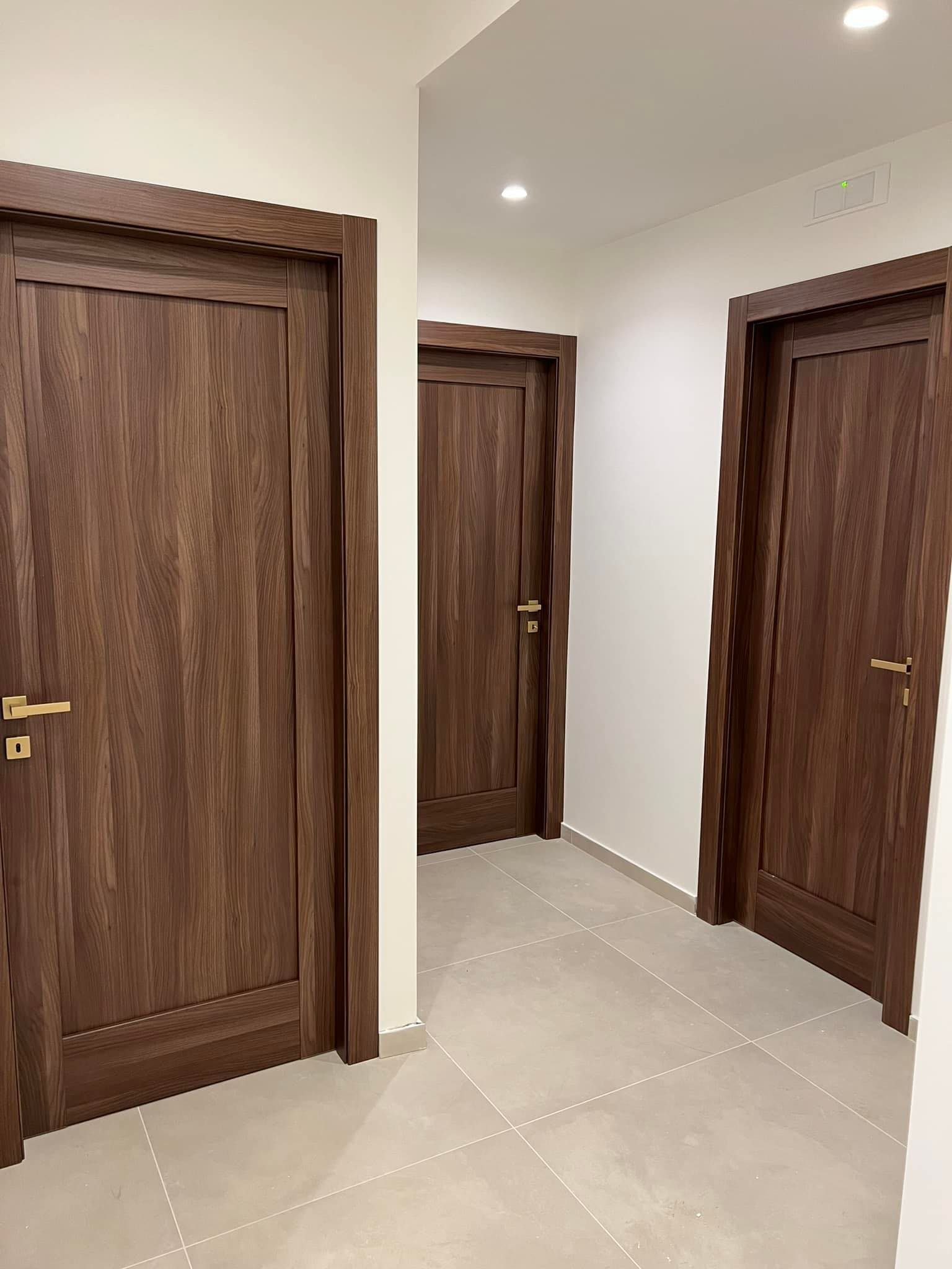 Tre porte di legno marrone in un corridoio con pavimento in piastrelle chiare e pareti bianche.