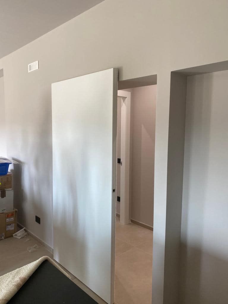 Porta scorrevole bianca parzialmente aperta, che rivela un corridoio. Pareti beige, illuminazione neutra.