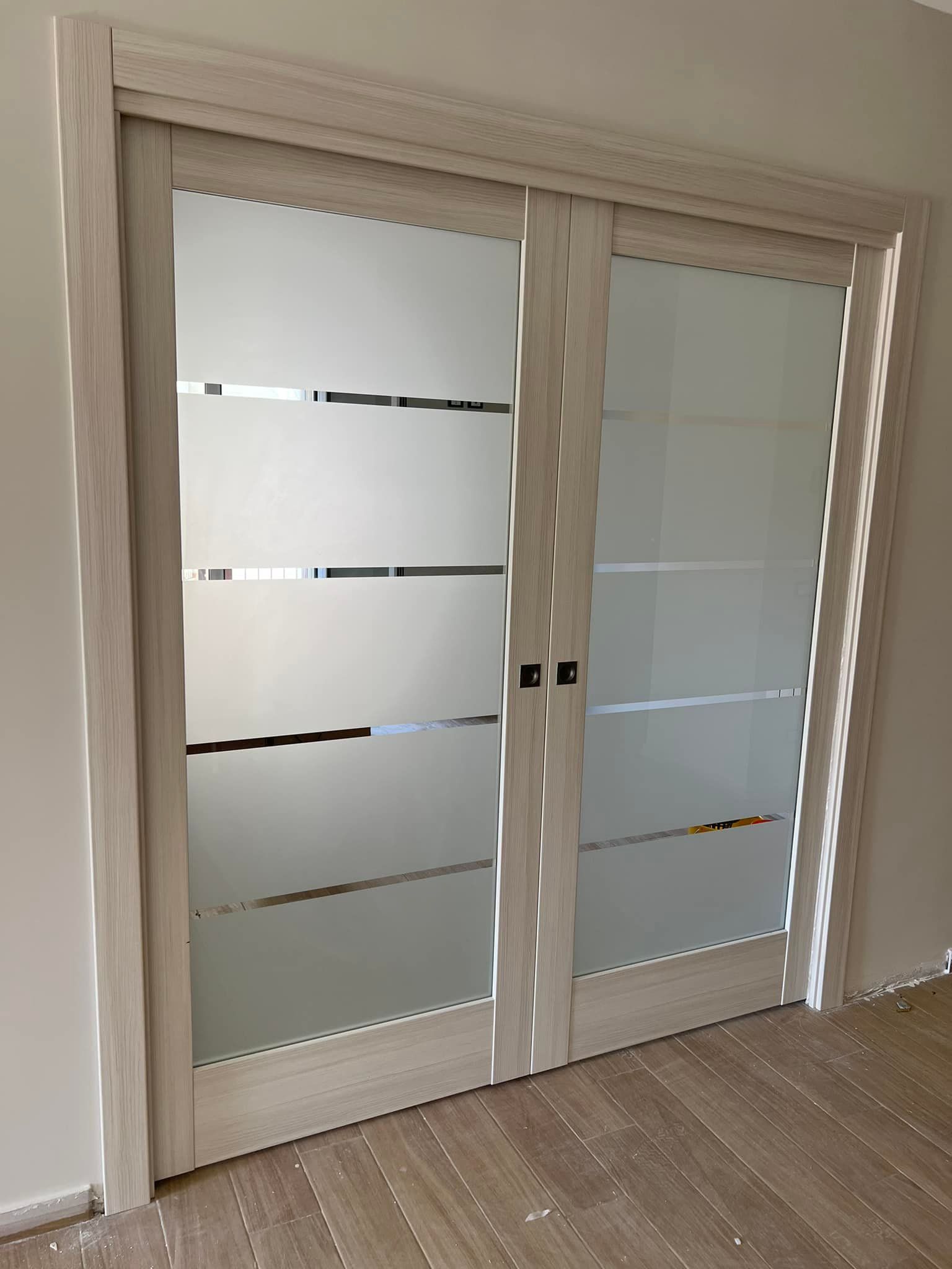 Doppie porte scorrevoli in vetro con pannelli in vetro satinato e strisce orizzontali satinate, con cornice in legno chiaro.