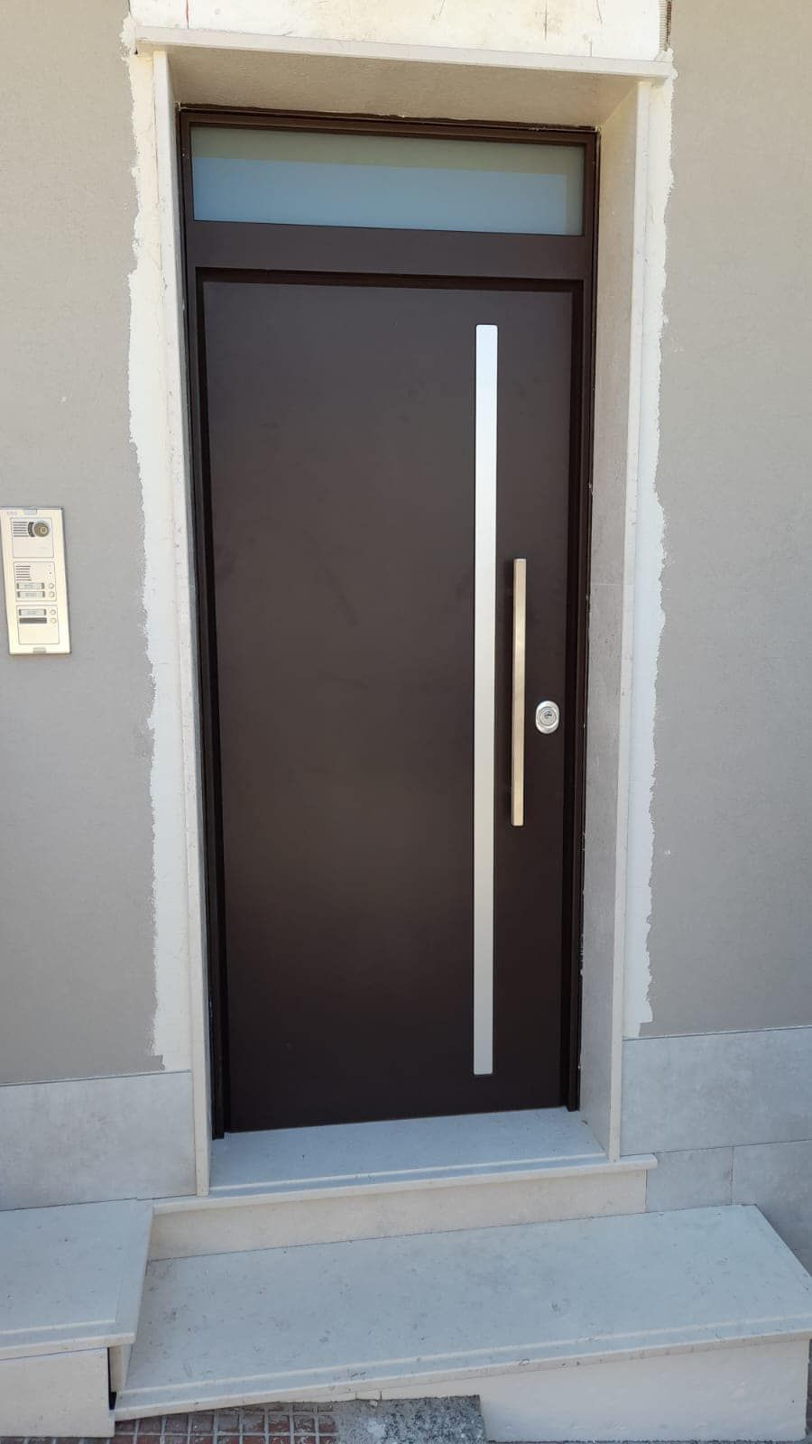 Porta d'ingresso nera con maniglia verticale argentata, finestra a traverso, inserita nella parete esterna grigia.