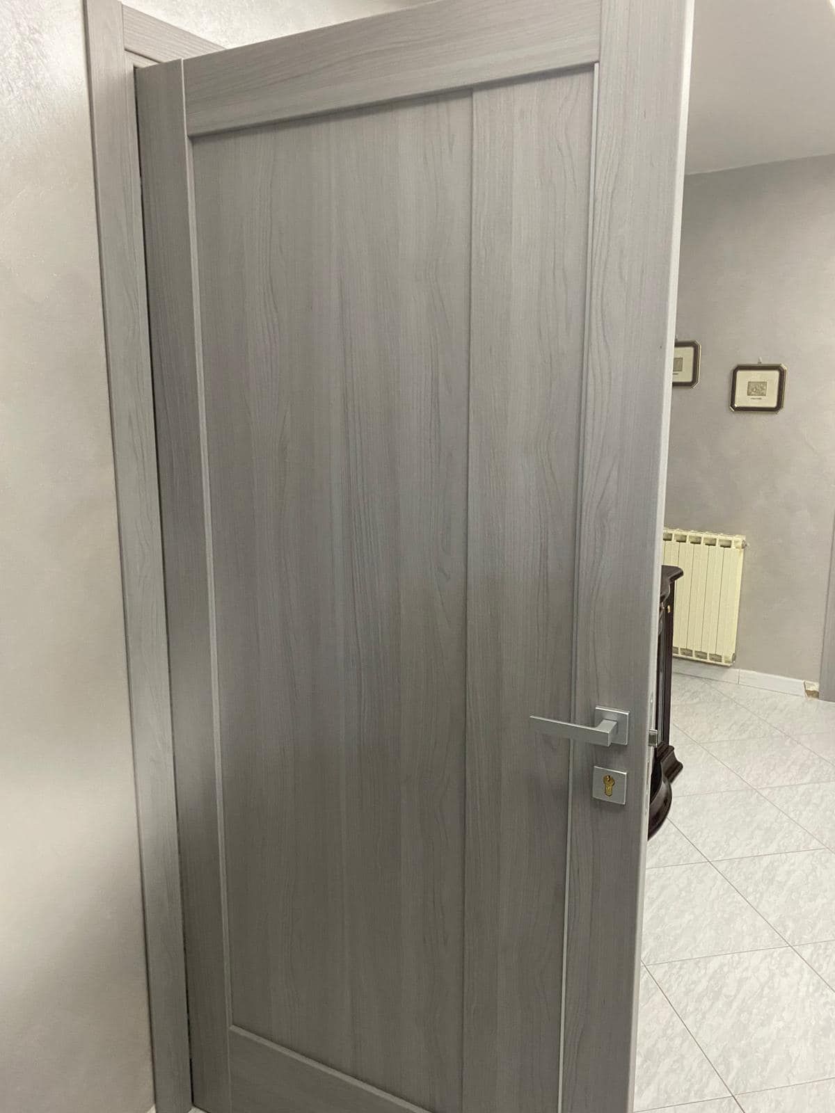 Porta interna in legno grigio, parzialmente aperta. È visibile una maniglia argentata, mentre lo stipite e la parete sono grigio chiaro.