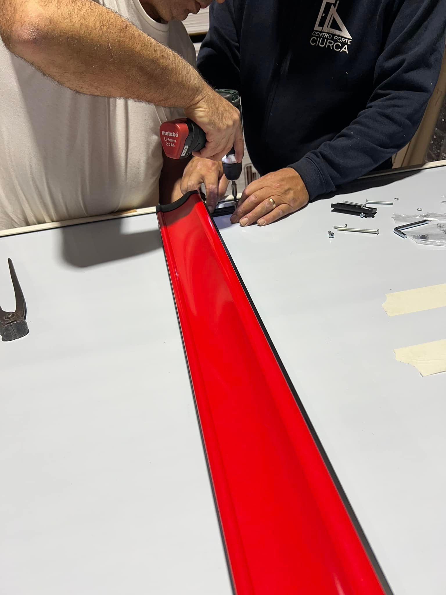 Due persone forano un pezzo di materiale rosso su una superficie grigia. Una persona tiene in mano il trapano.