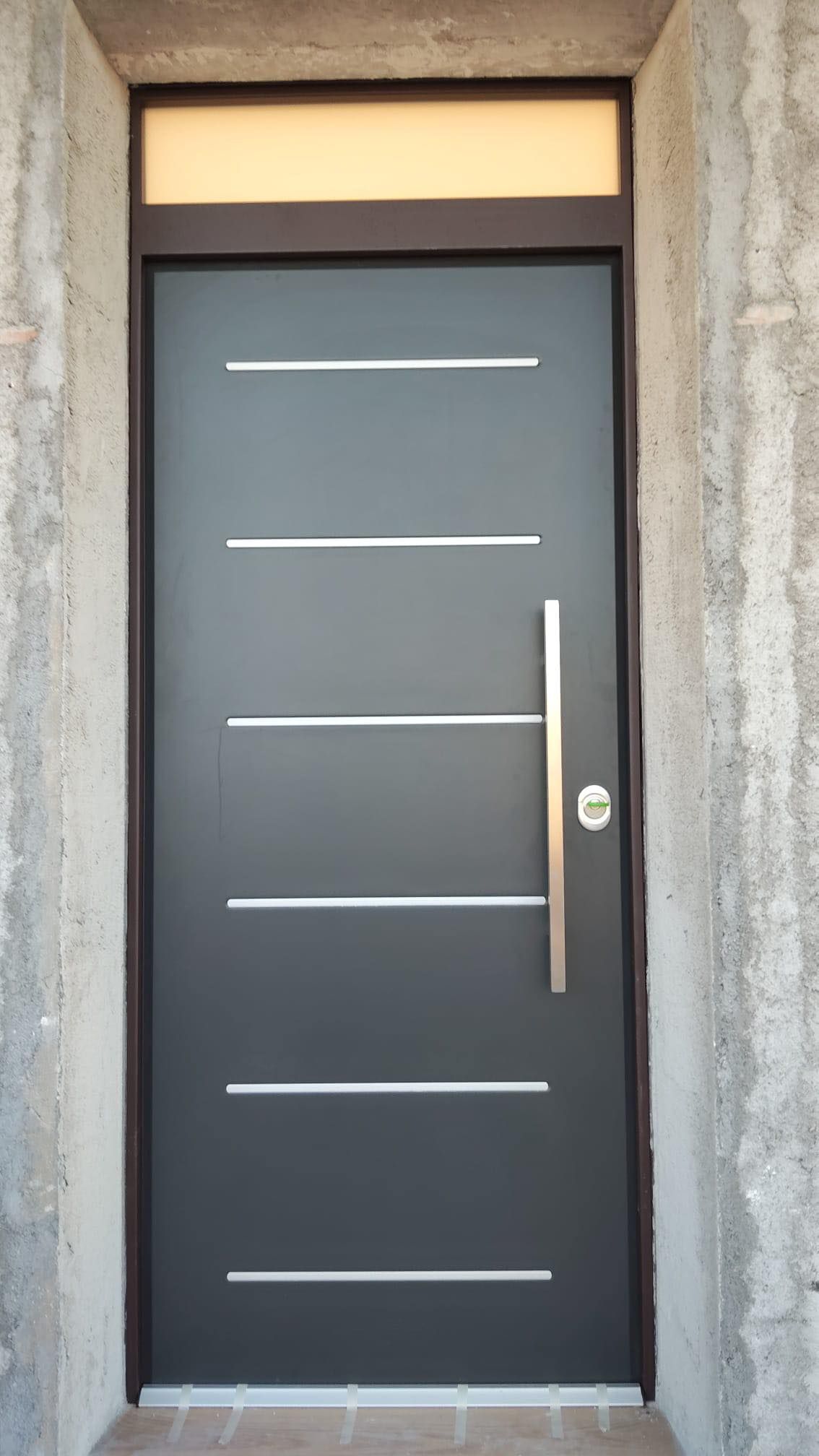 Porta d'ingresso grigio scuro con linee orizzontali argentate e maniglia verticale. Sopra c'è una finestra.