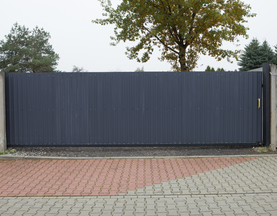 Cancello scorrevole grigio scuro su un vialetto in mattoni e cemento, con alberi sullo sfondo.