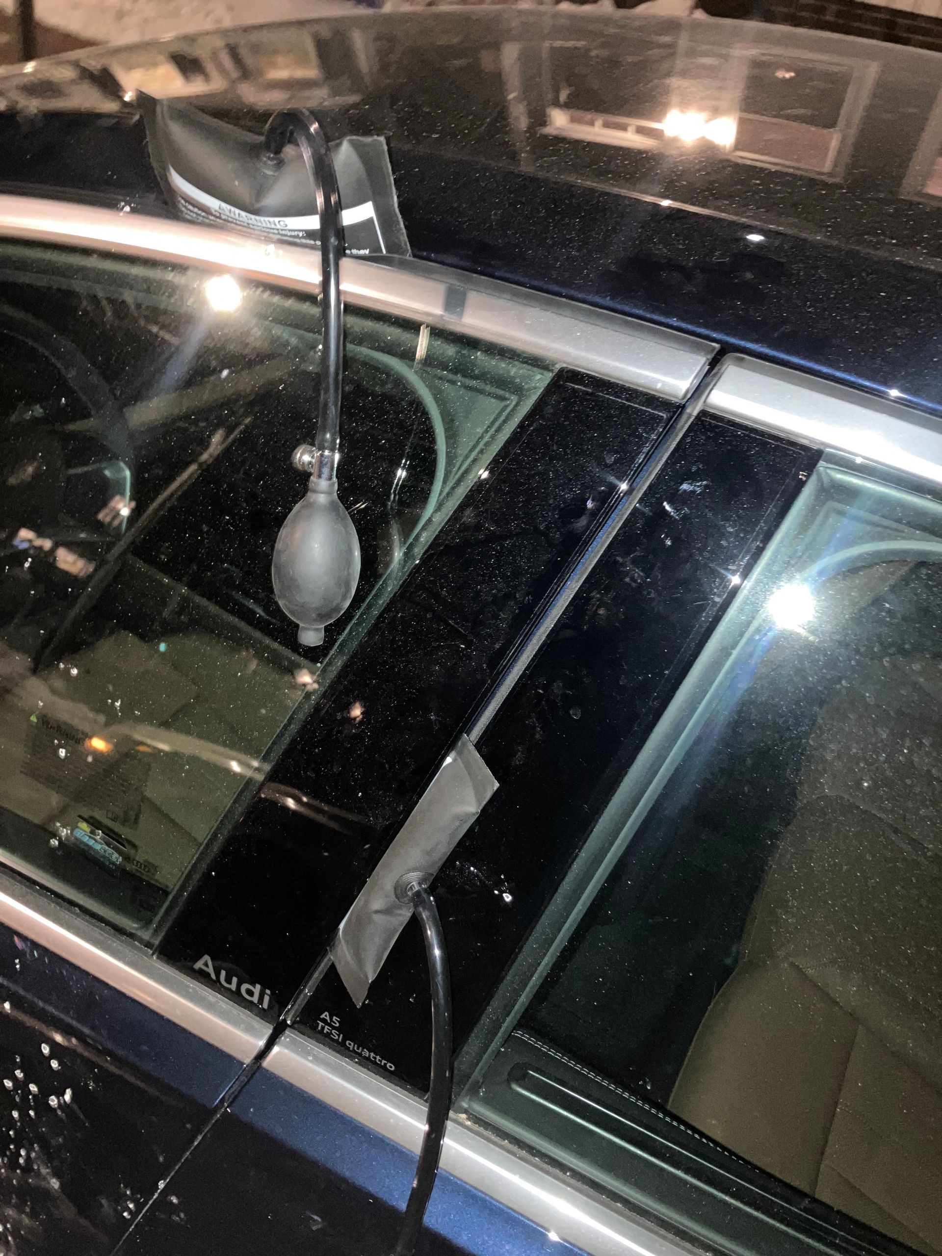 Broken car window with a dangling rearview mirror and visible shards on a dark vehicle.