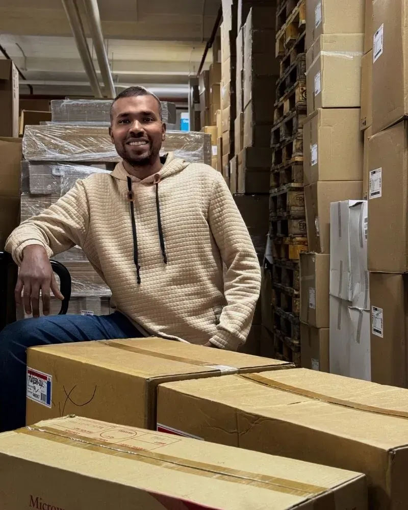 Ein Mann sitzt lächelnd in einem Lagerhaus zwischen Kisten. Im Hintergrund sind Reihen gestapelter Kisten zu sehen.