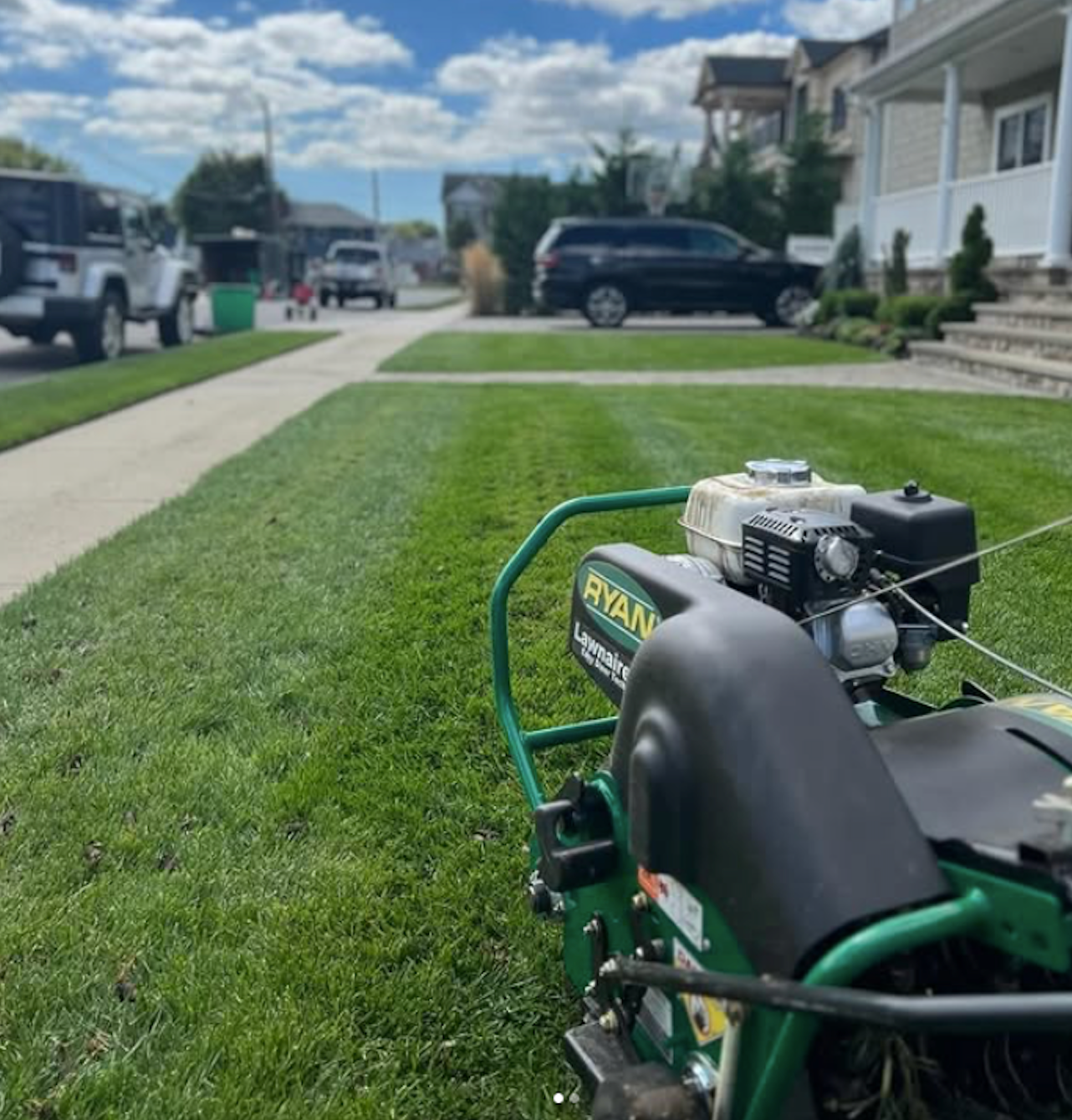 Green lawnmower on a suburban sidewalk beside a neatly cut lawn and houses on a sunny street