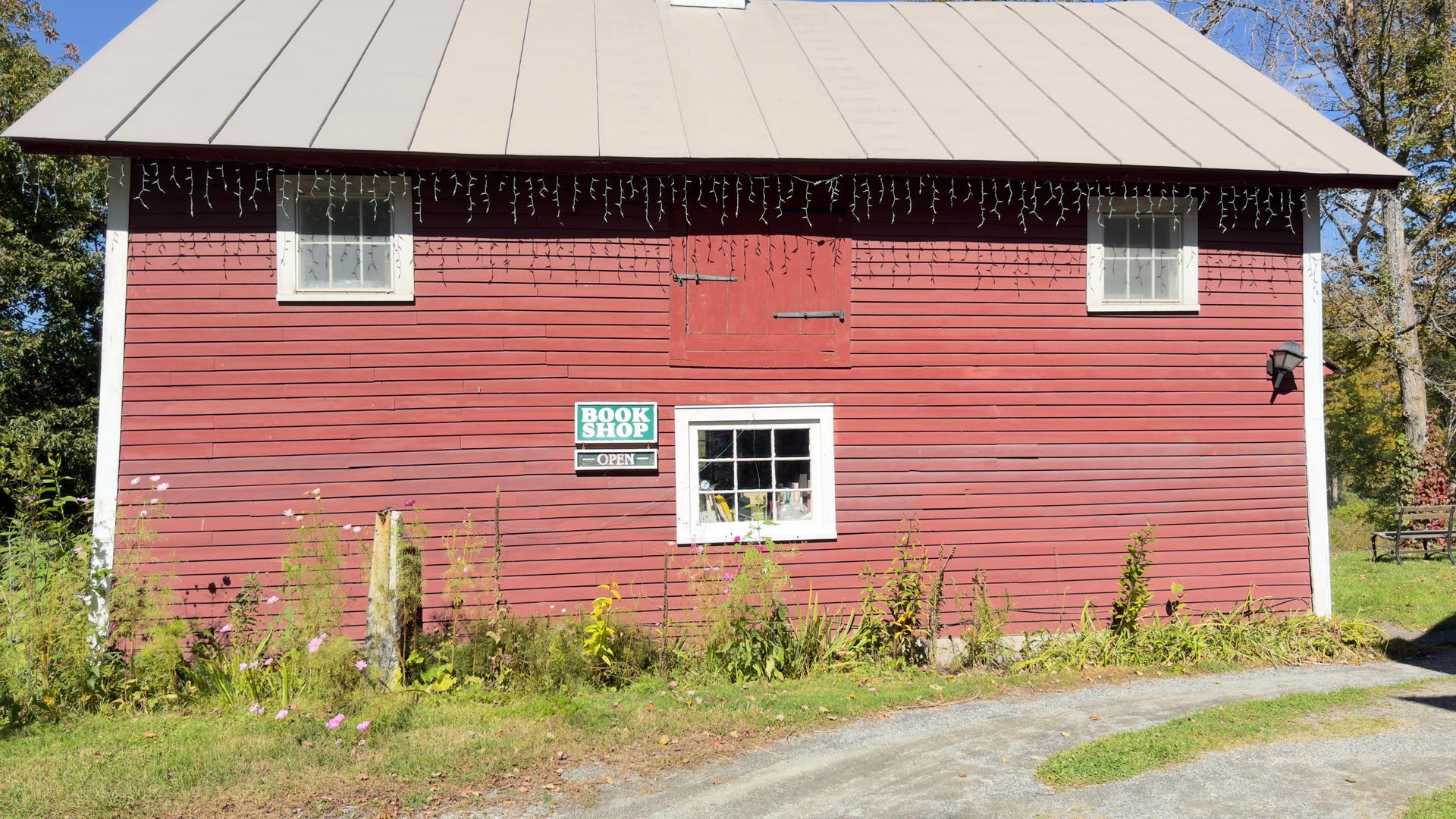 Red barn with a tan metal roof, small white windows, and overgrown grass beside a gravel drive.