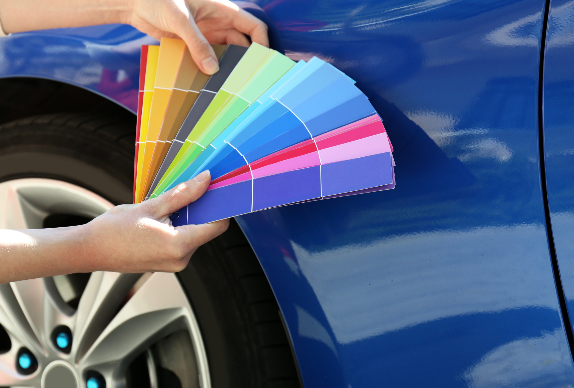 Hands holding a color palette fan against a blue car, comparing paint shades.