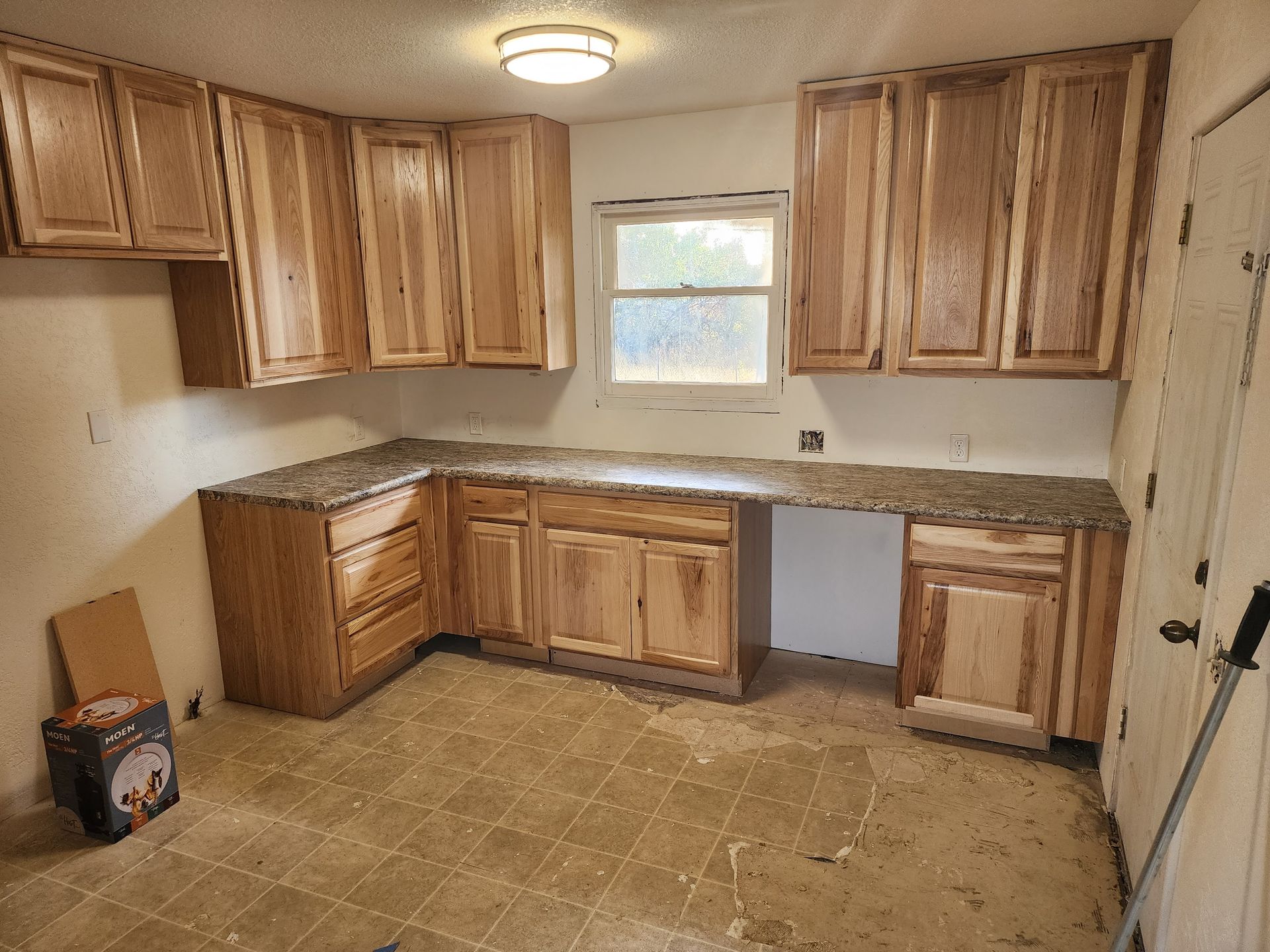 Kitchen with light wood cabinets, speckled countertop, small window, and worn flooring.