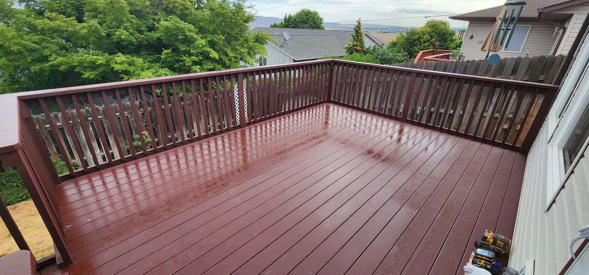 A wooden deck with brown planks and railings, wet from rain, surrounded by green trees and houses.