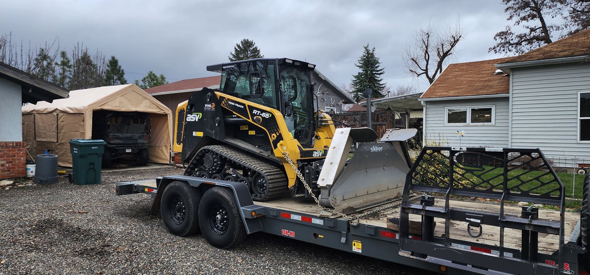 A yellow and black skid steer loader on a trailer parked in a residential driveway.