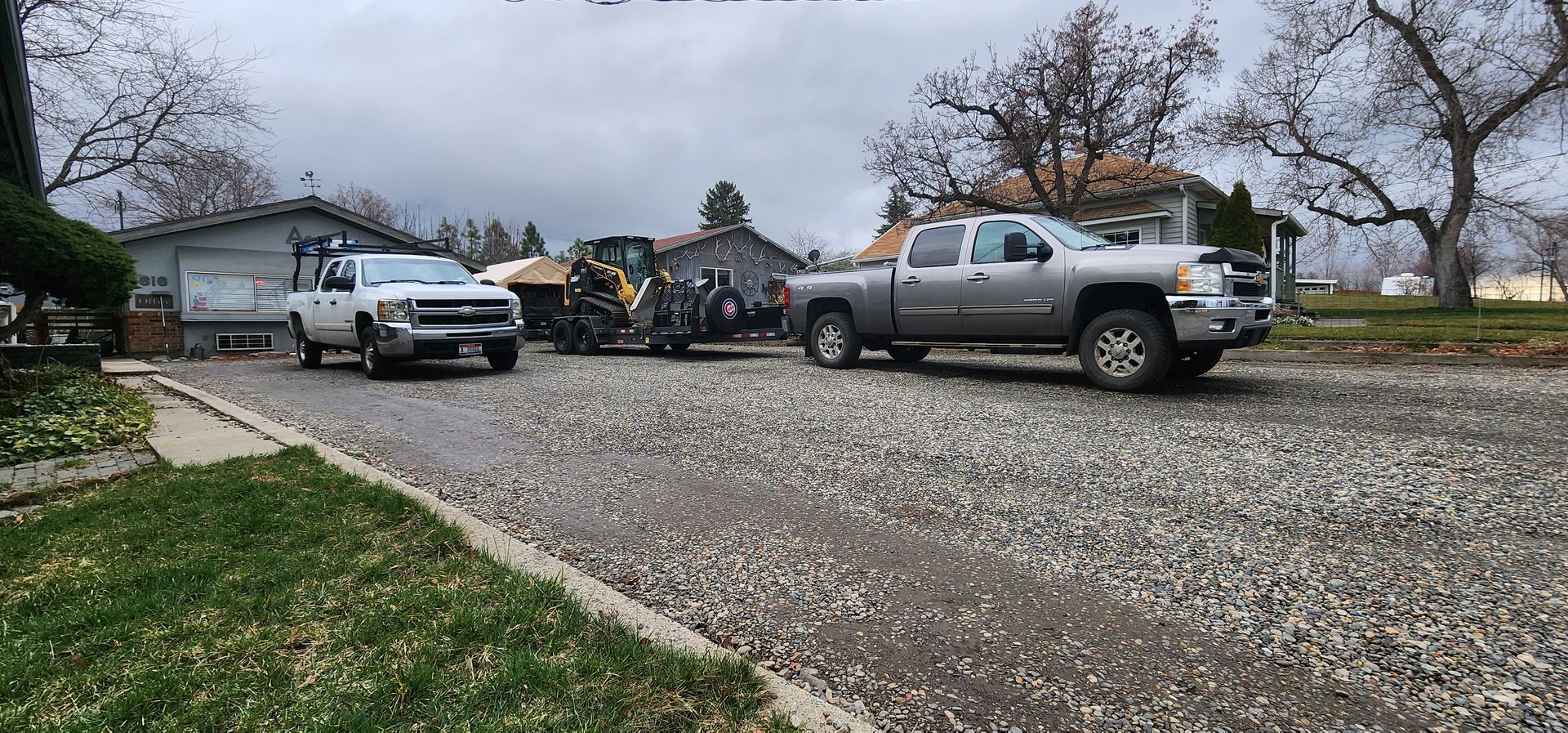 Three trucks parked on a gravel driveway in front of a house on a cloudy day.