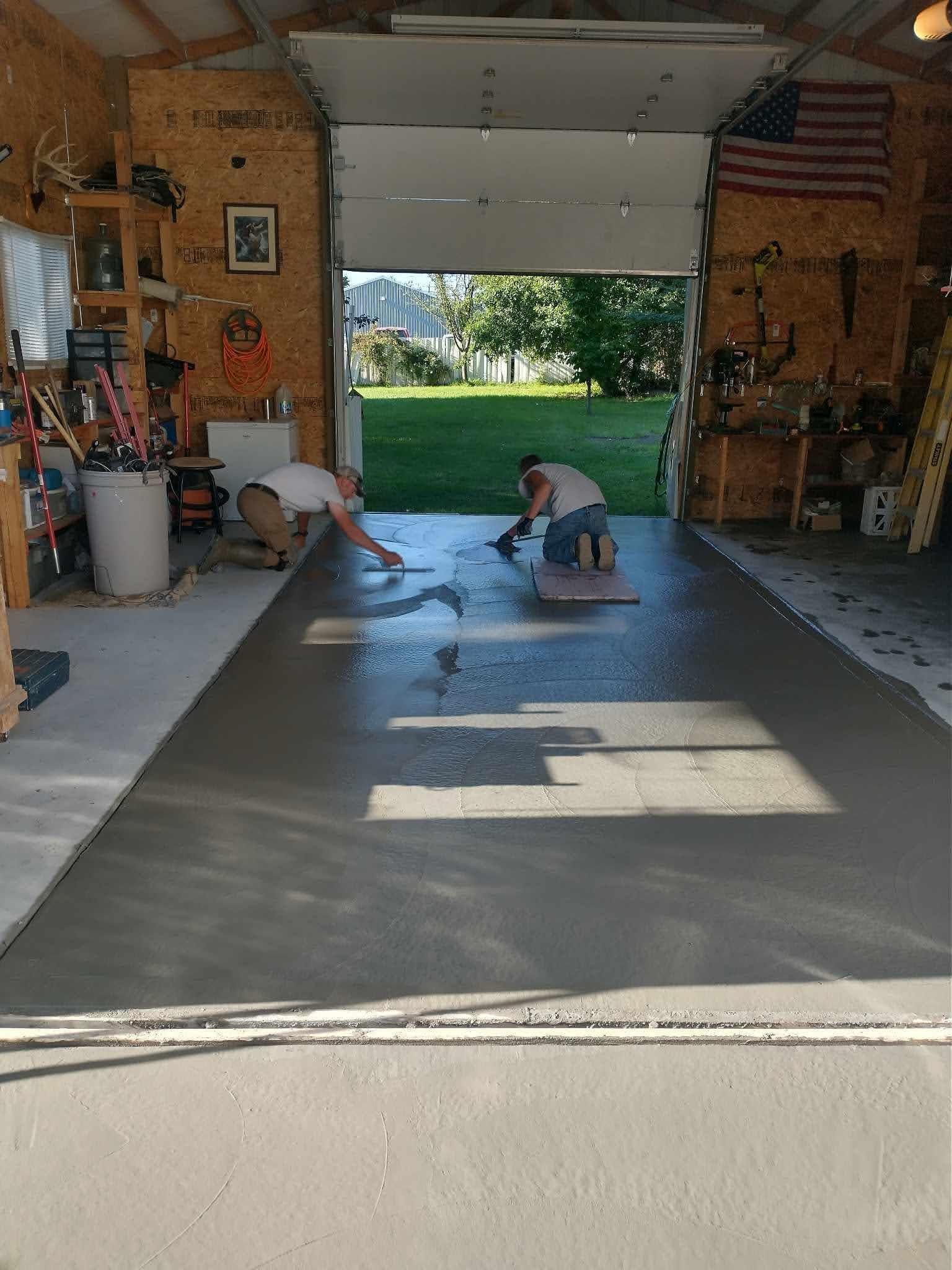 Two people smoothing wet concrete in an open garage. Green lawn visible through the doorway.