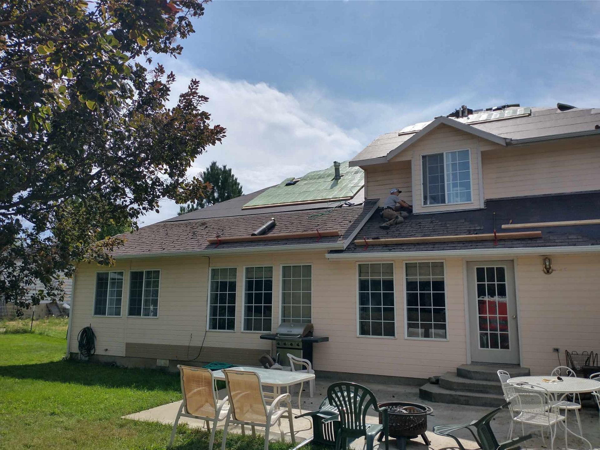 Back of a two-story beige house with roof repair underway, patio furniture, and a tree on a sunny day.