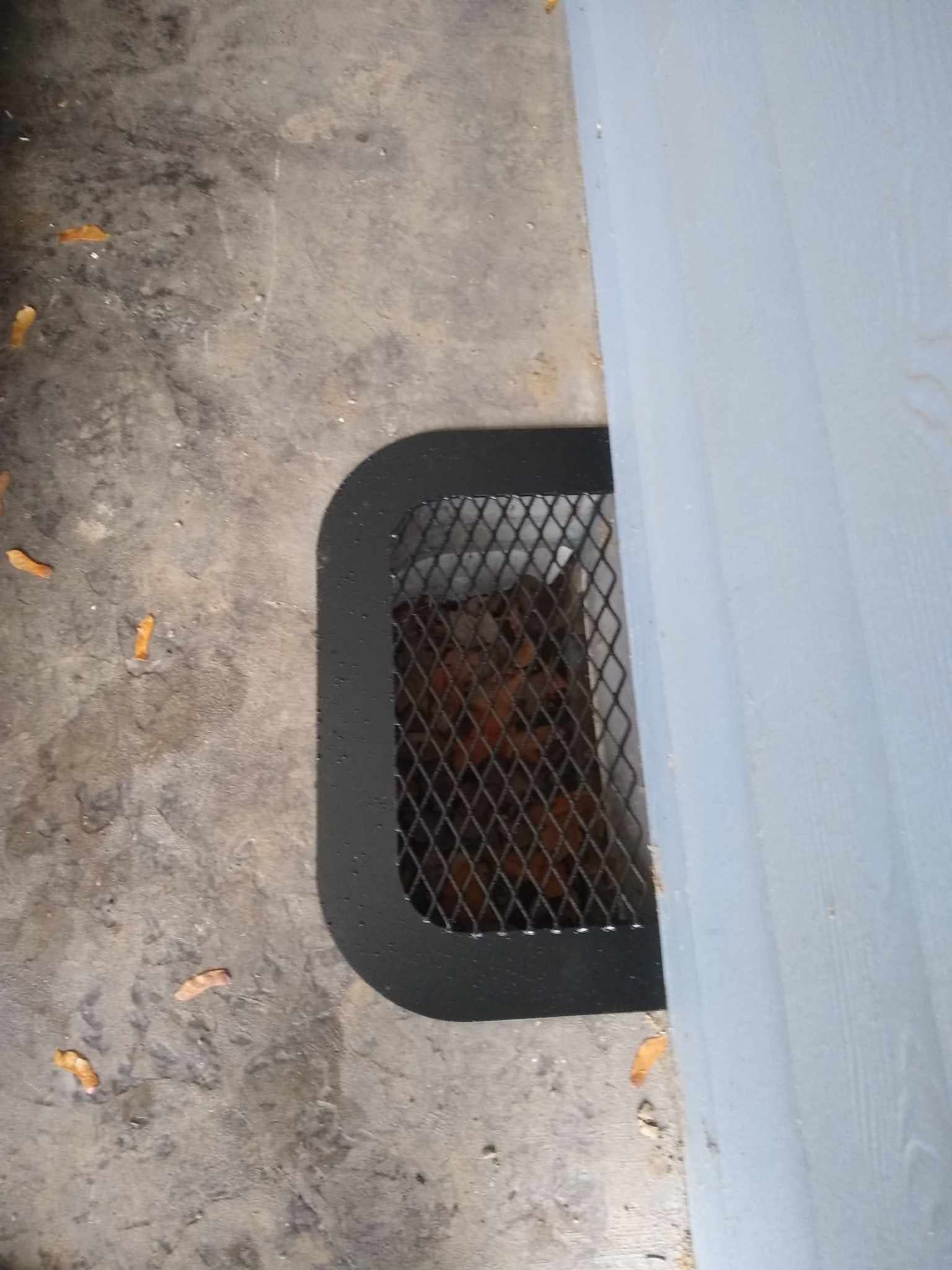 Black metal grate covering a recessed opening in concrete, next to a light blue wall.