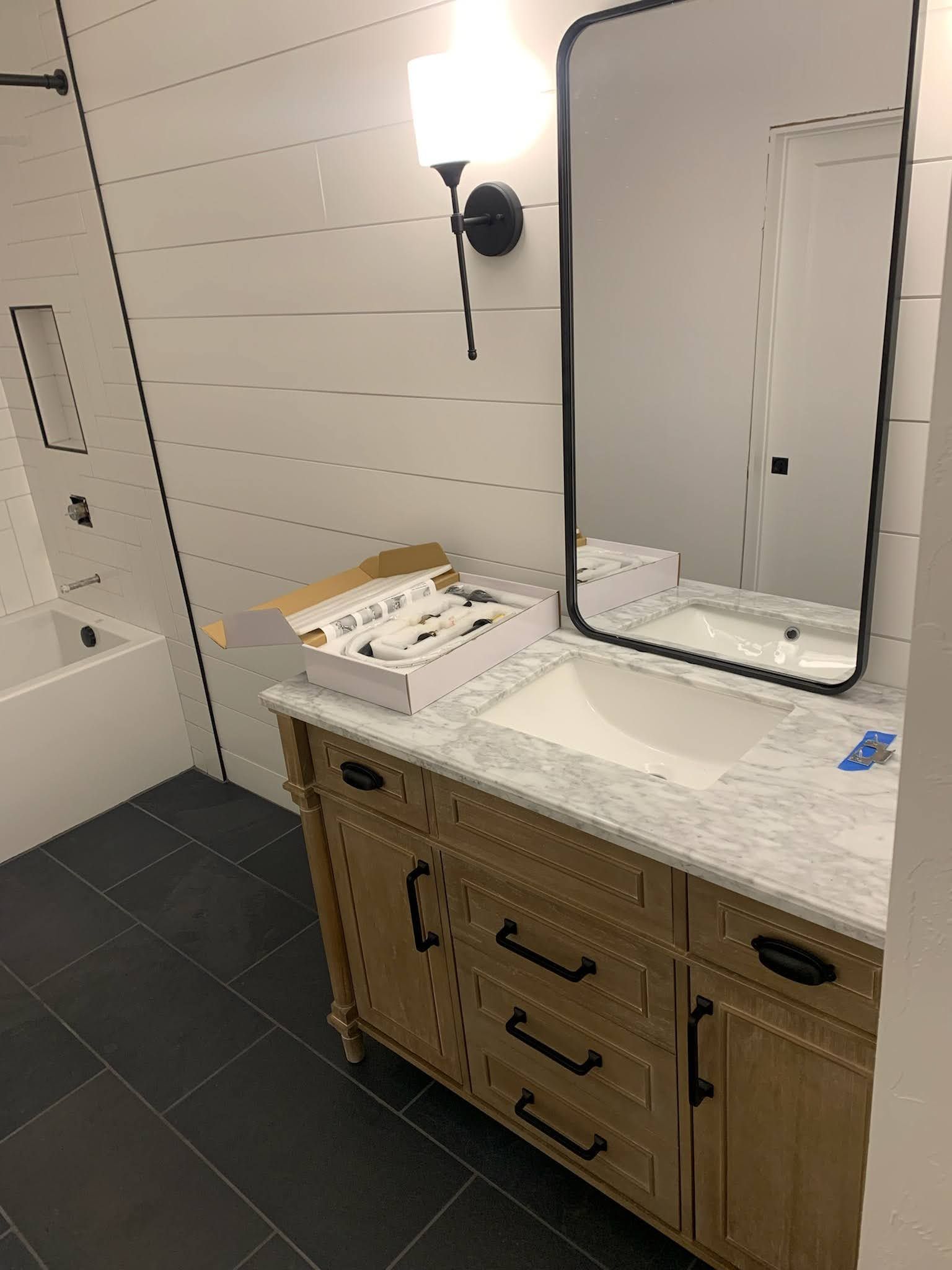 Bathroom with light-colored vanity, white sink, black-framed mirror, and white shiplap walls. Black tile floor.
