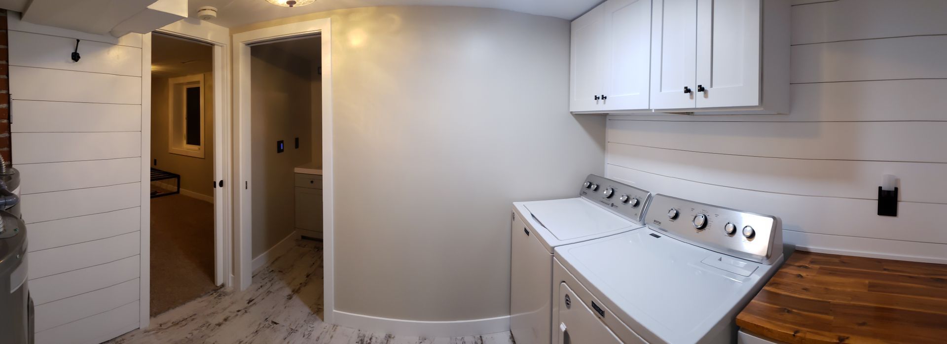 Laundry room with white cabinets, appliances, and shiplap walls. Wooden countertop, two doorways in view.