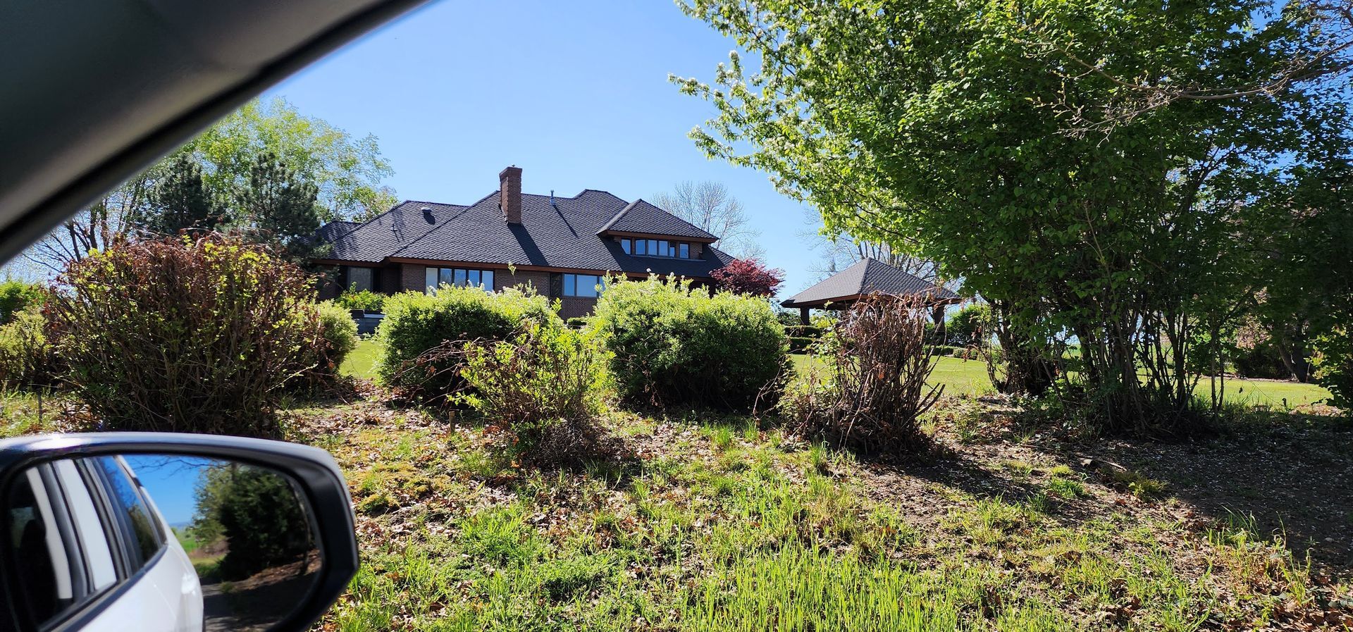 View of a large house and gazebo through a car window. Sunny day, green foliage.